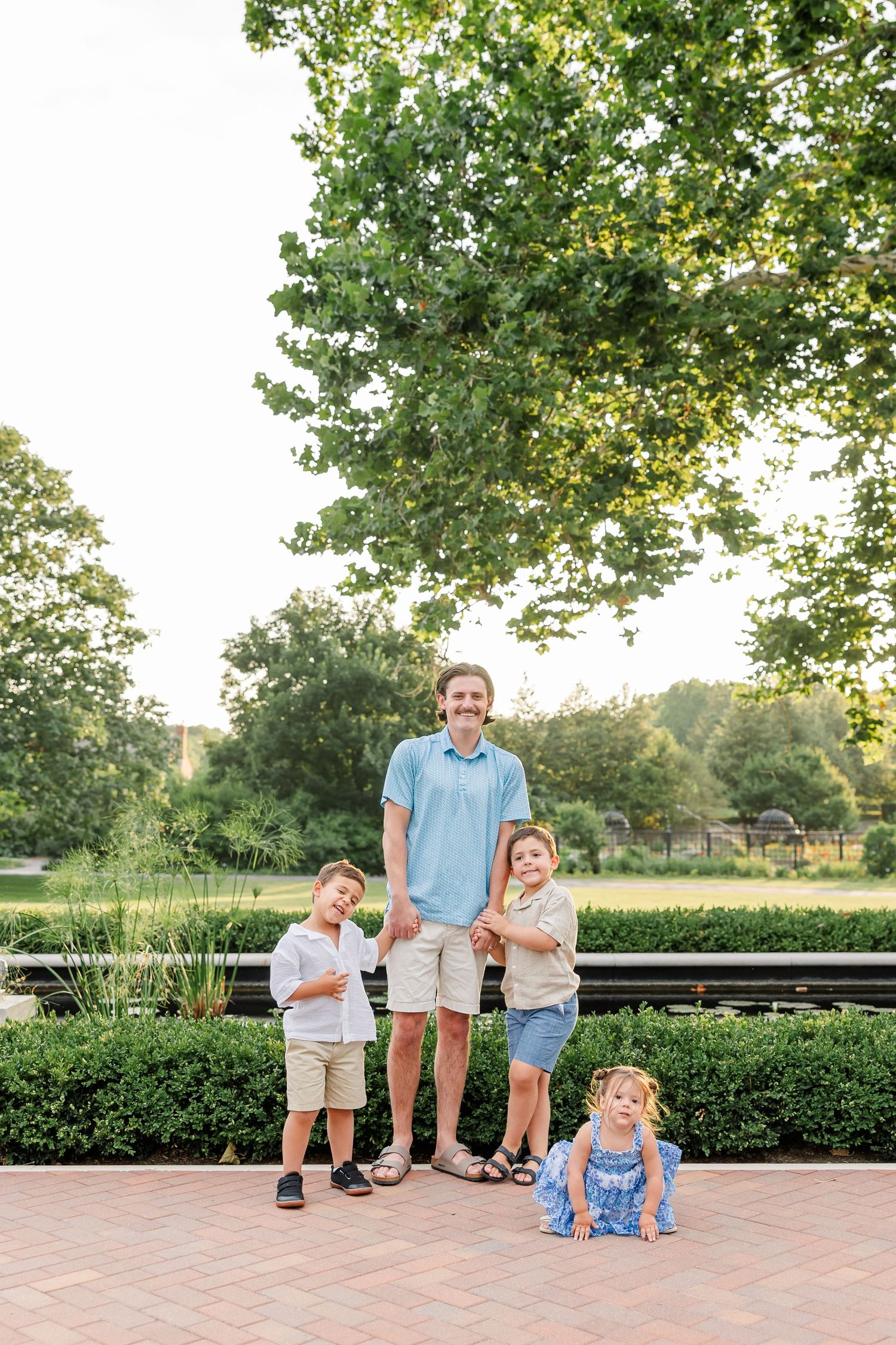 Father posing with three young children on a brick walkway in a sunny green park setting.