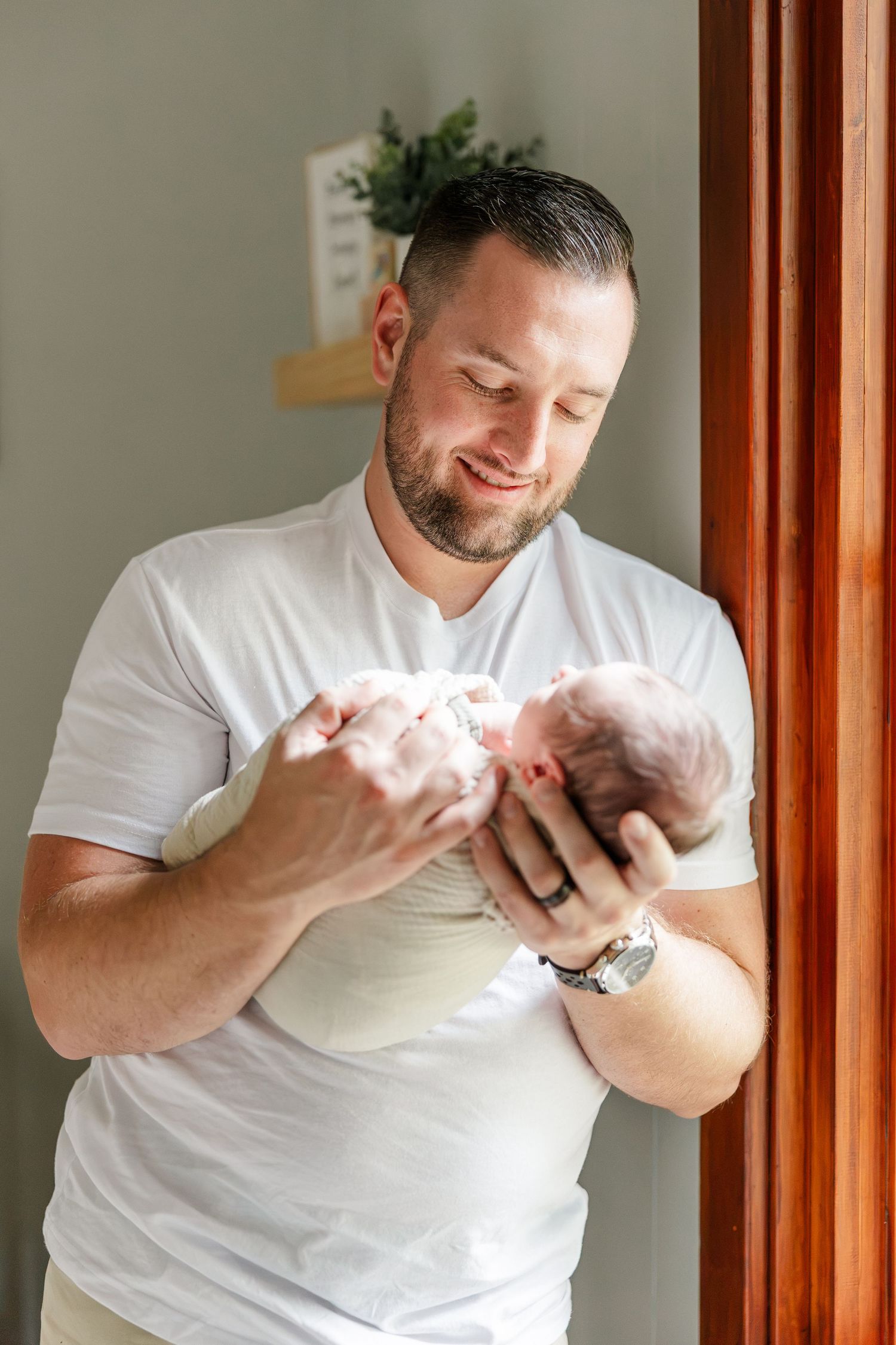 Smiling father in white t-shirt tenderly holds newborn baby near a window in a warmly lit room.