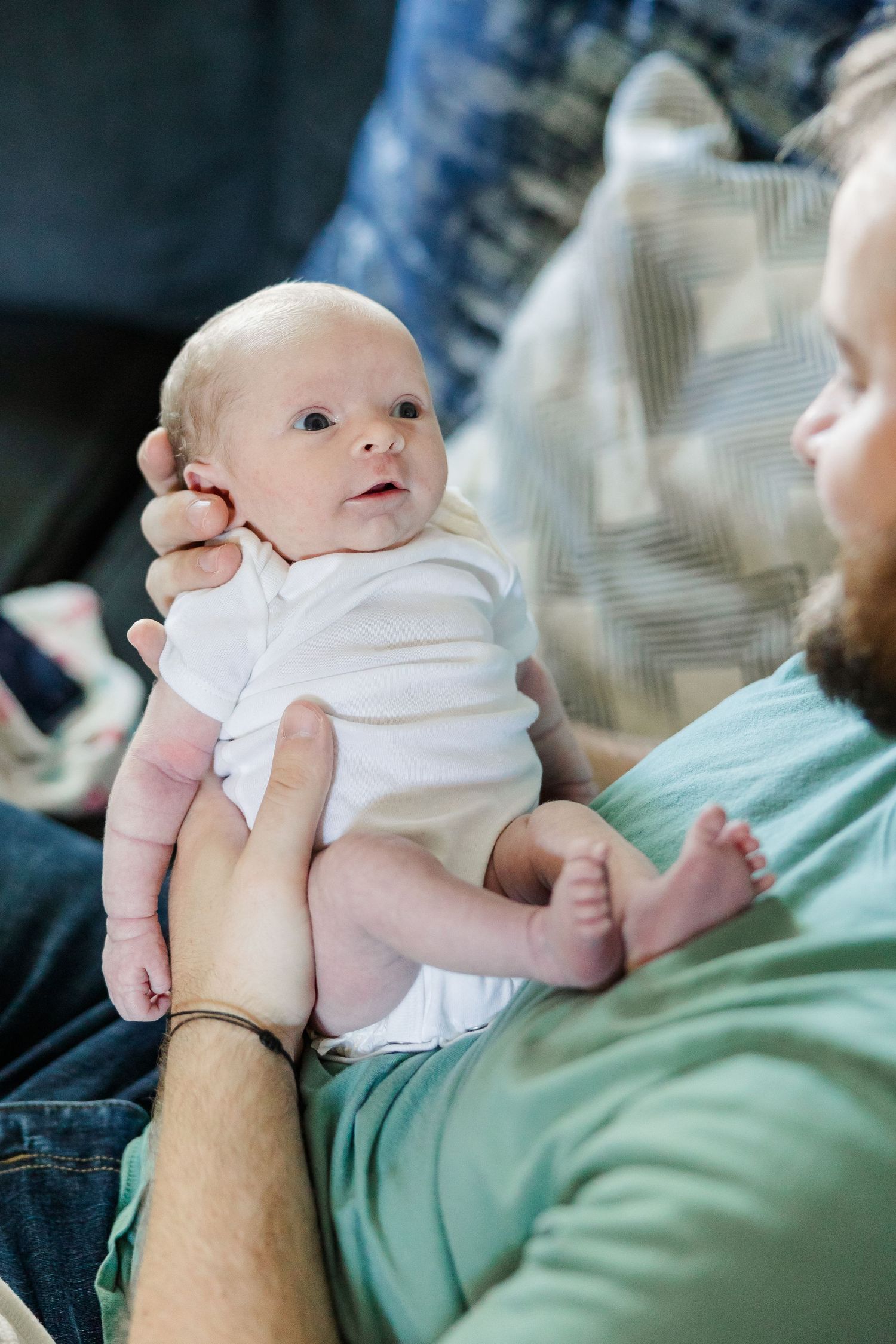 Father holding newborn baby in white onesie while sitting on couch at home.