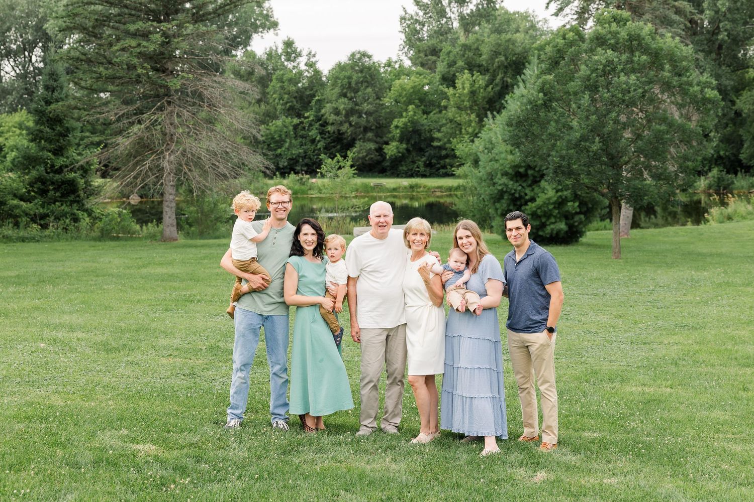 Multi-generational family of seven smiling together outdoors on green lawn with lush trees in background.