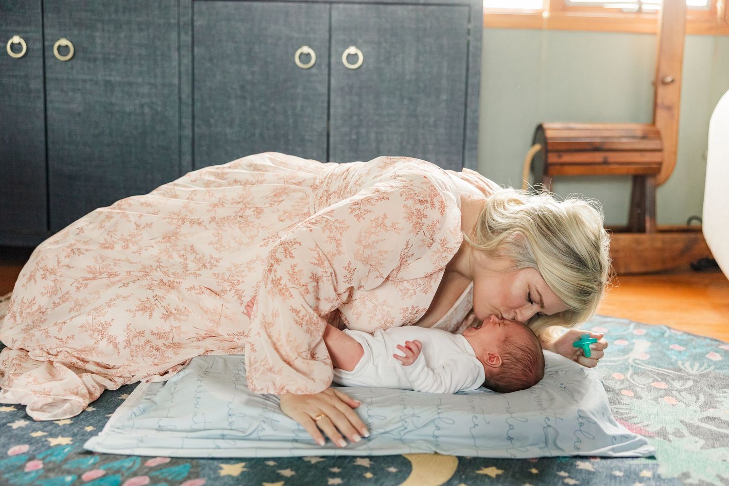 Loving mother leaning down to kiss her newborn baby lying on a play mat on the floor.