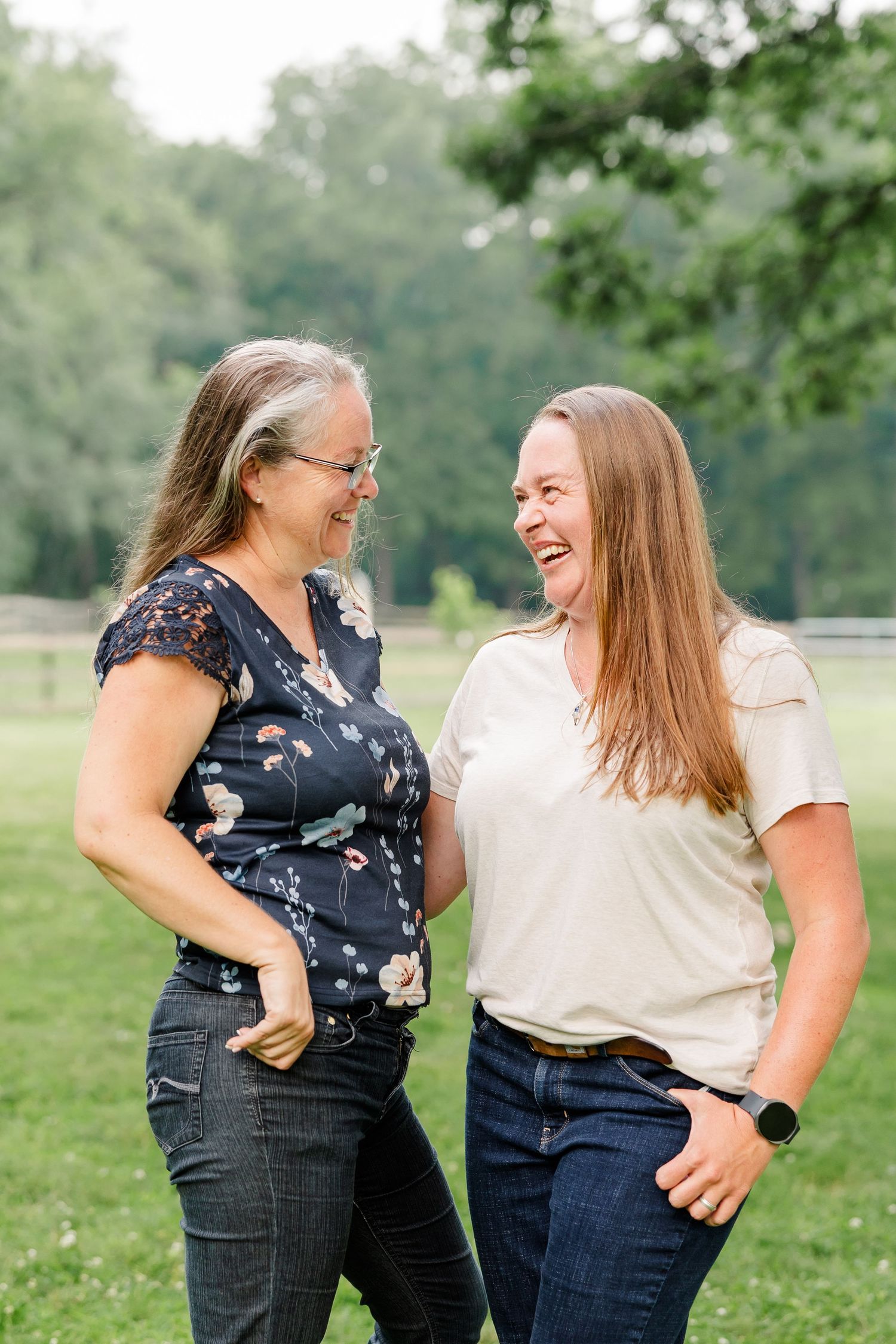 Two women laughing together outdoors in a green park setting, one wearing a floral blouse and glasses.