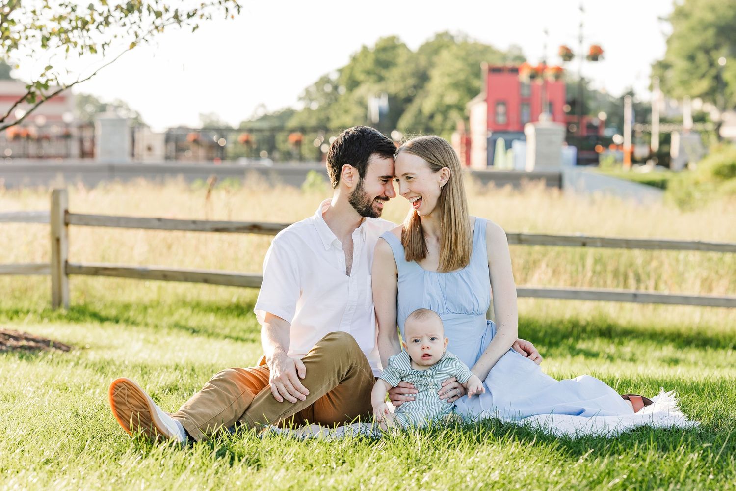 Young couple sitting on grass with baby in sunny park by wooden fence.