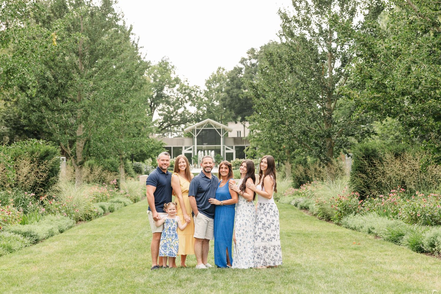 A joyful family of seven poses together on a lush garden path with trees and a gazebo backdrop.