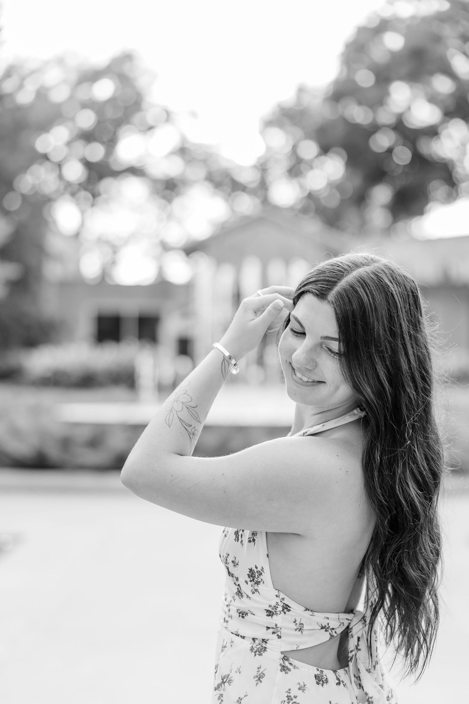 Black and white portrait of a smiling young woman in a floral dress, posing outdoors with her hand in her hair.