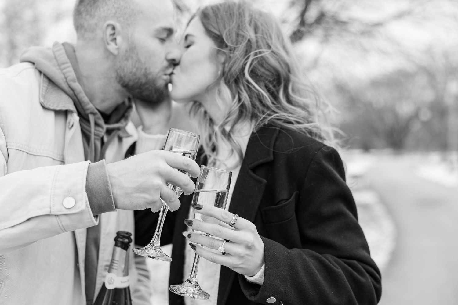 A couple shares a romantic kiss while holding champagne glasses outdoors in black and white photography. Proposal photos in Winnemac Park,