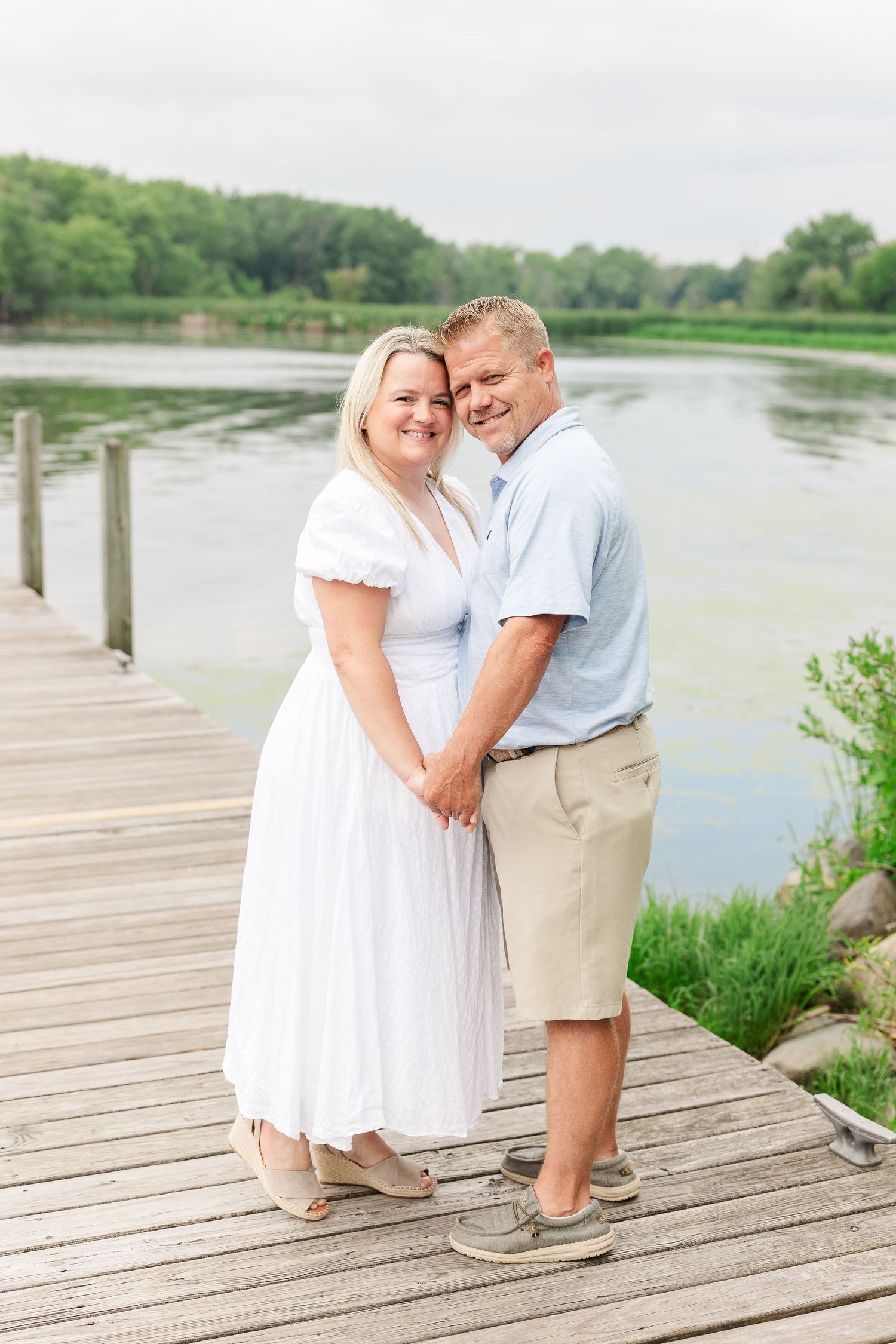 Couple in coordinated white and khaki outfits embrace on lakeside dock during summer photoshoot.