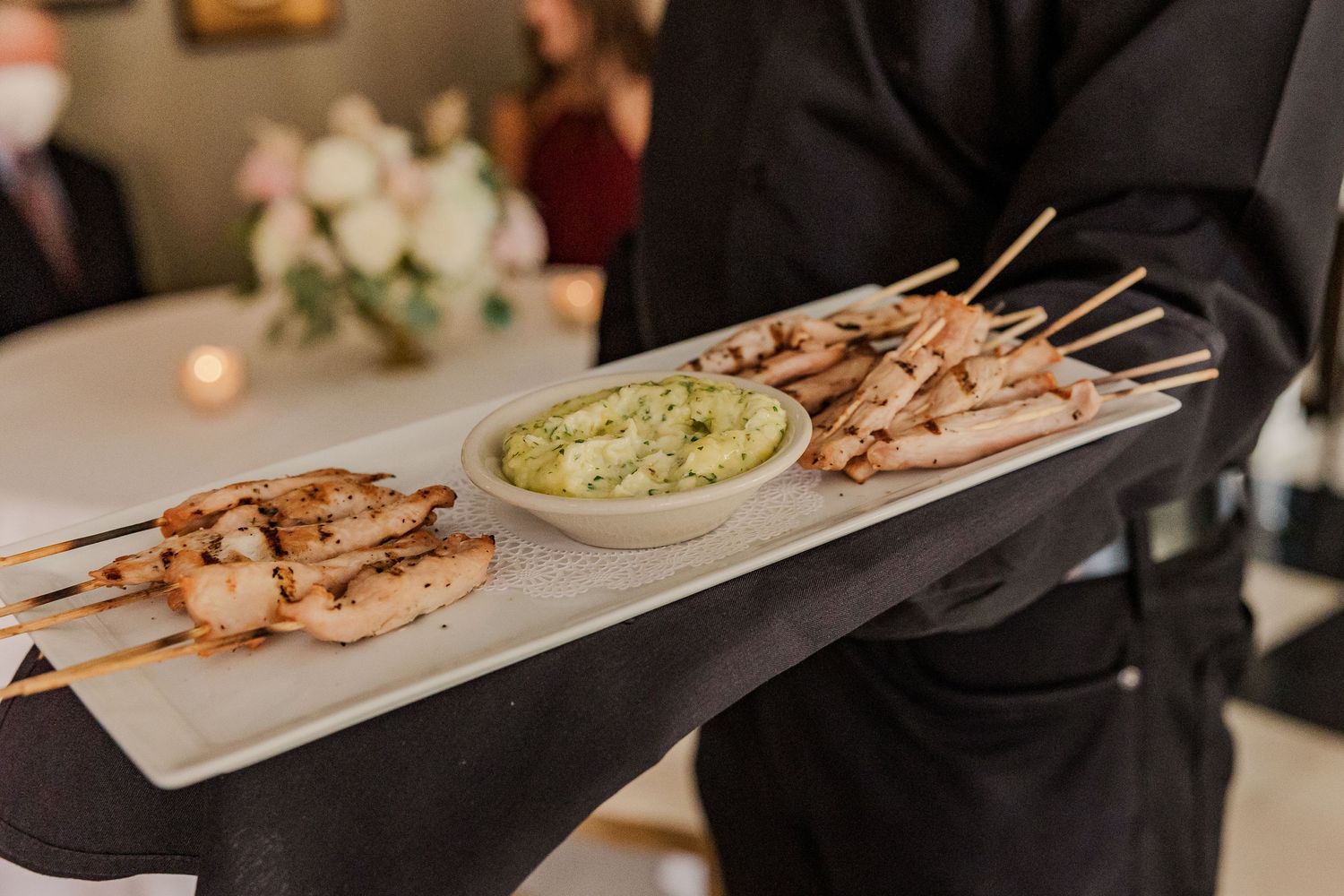 Catering server holding tray of grilled shrimp skewers with green dipping sauce.