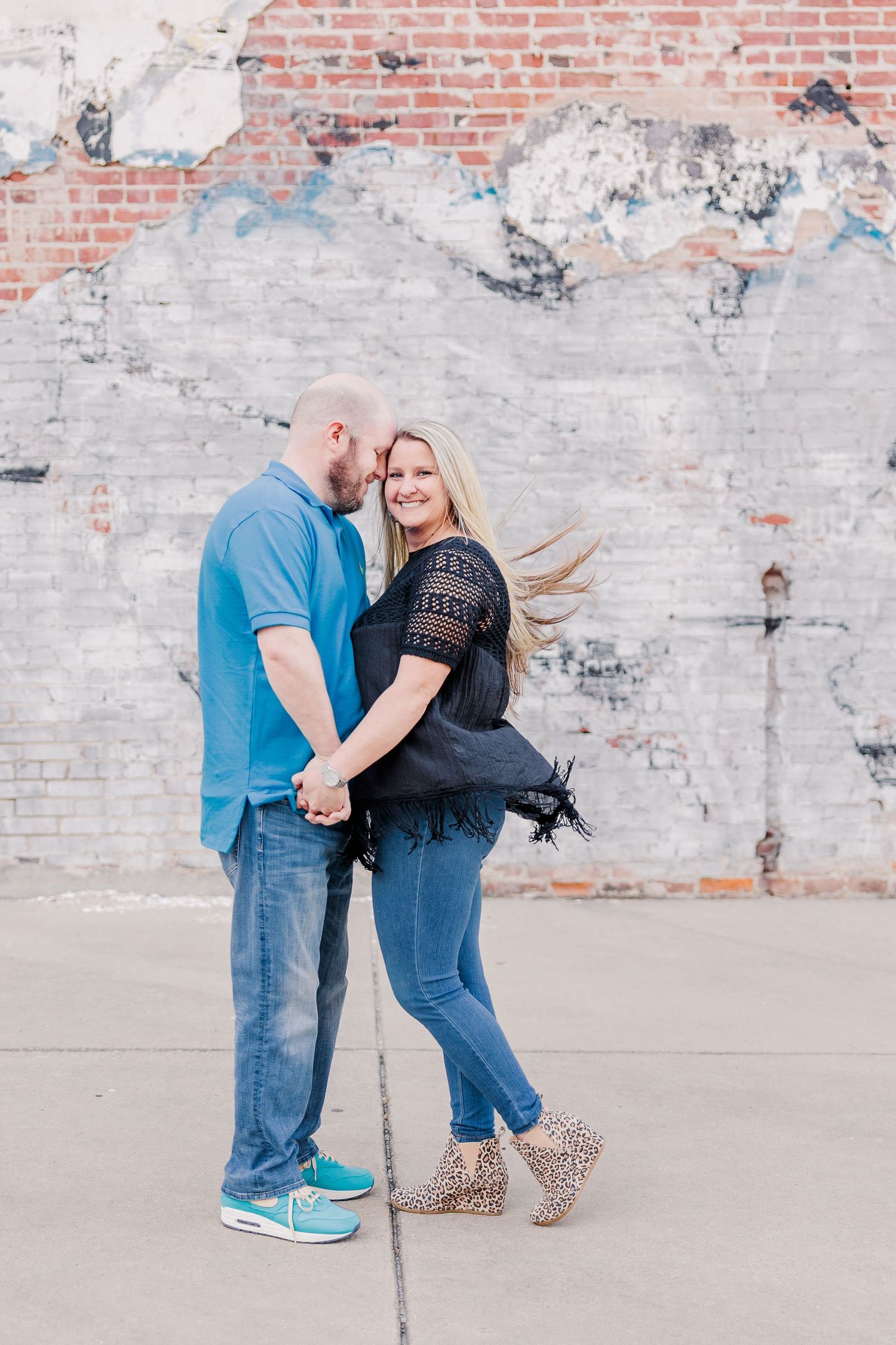 A couple embraces playfully in front of a distressed brick and concrete wall.