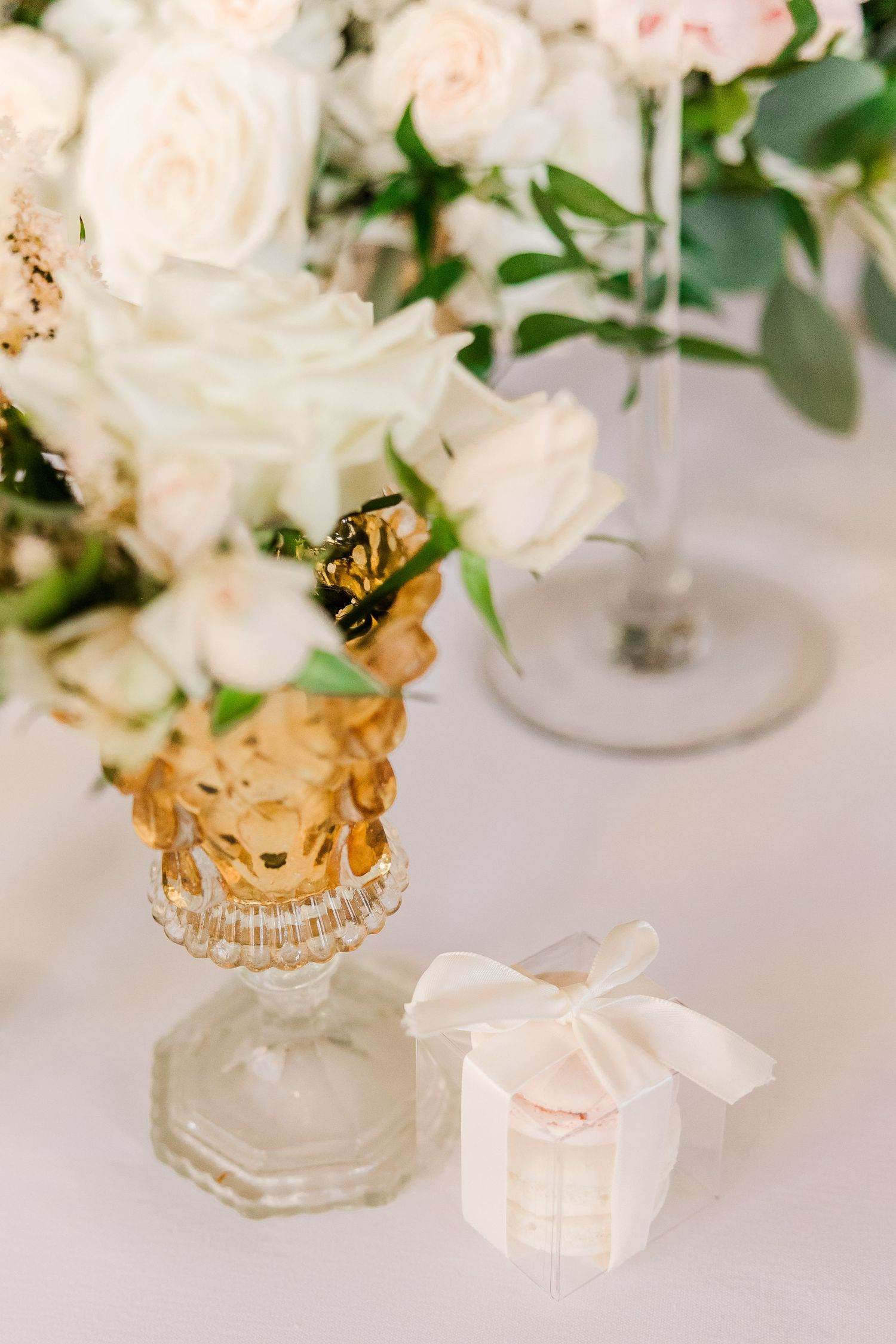 White roses arranged in a vintage gold-tinted glass vase with a small gift box on white tablecloth.
