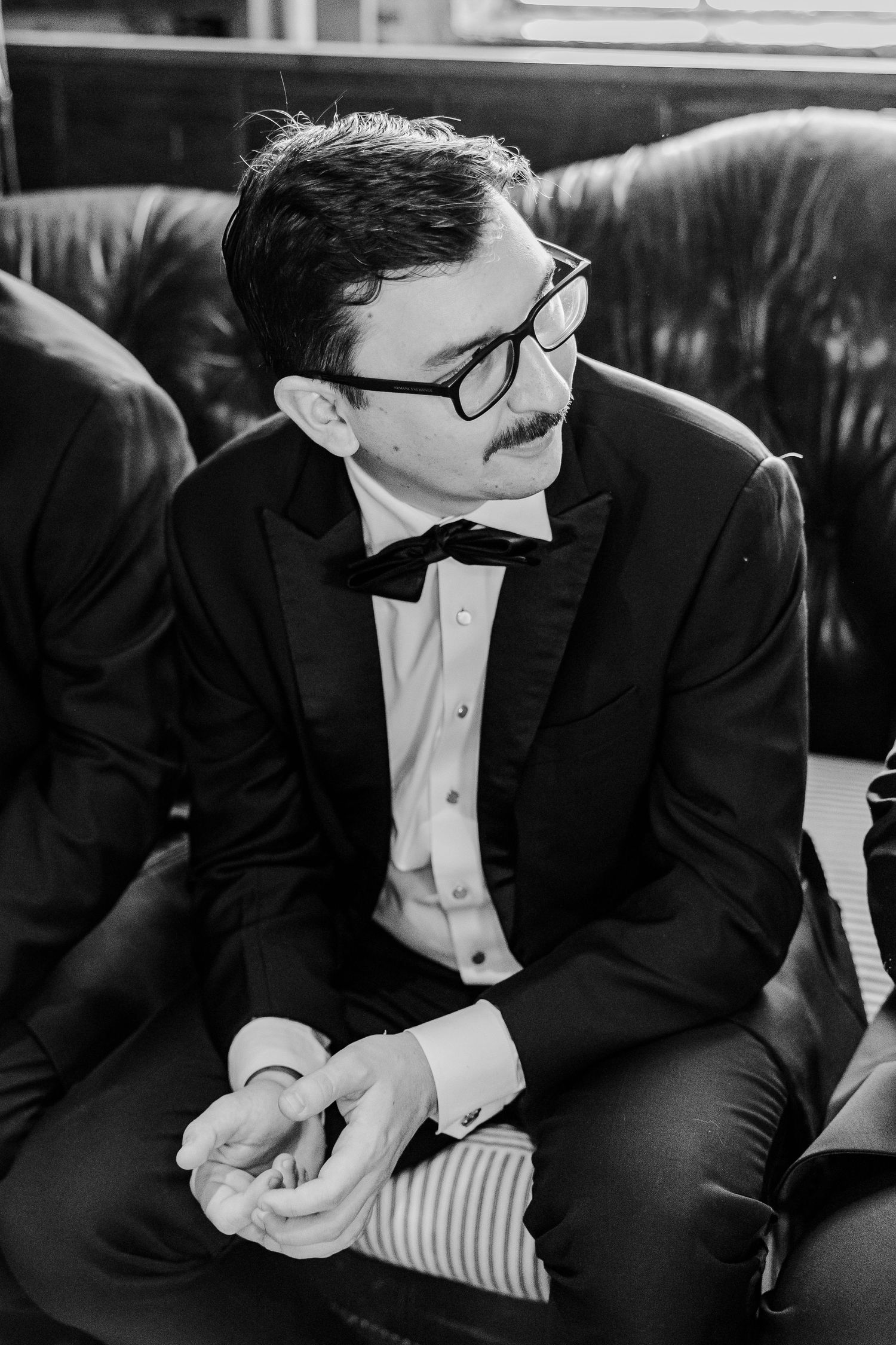 A groomsman in glasses and formal attire sits on a leather couch.