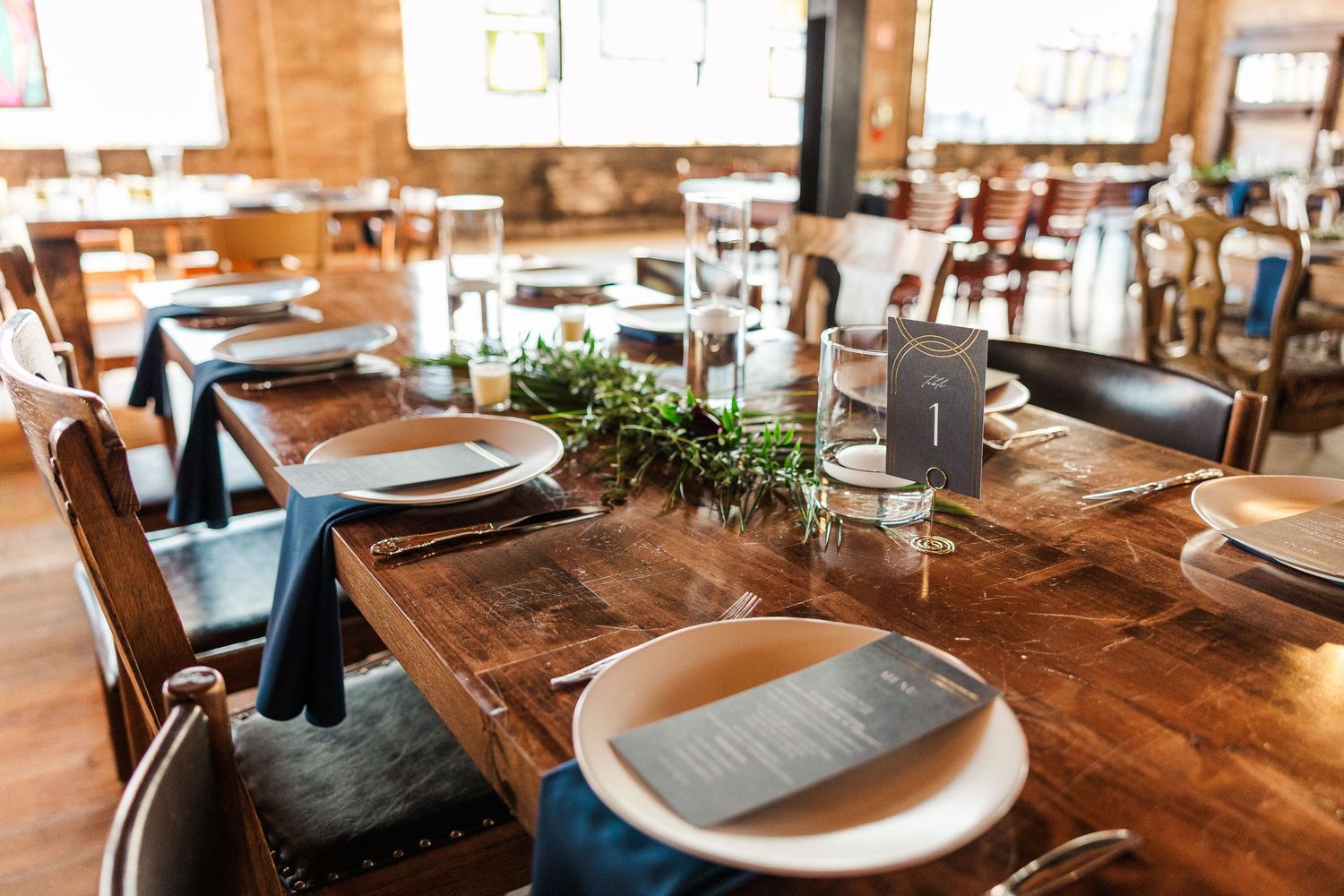 Rustic wooden dining tables set with plates and greenery centerpieces in restaurant.