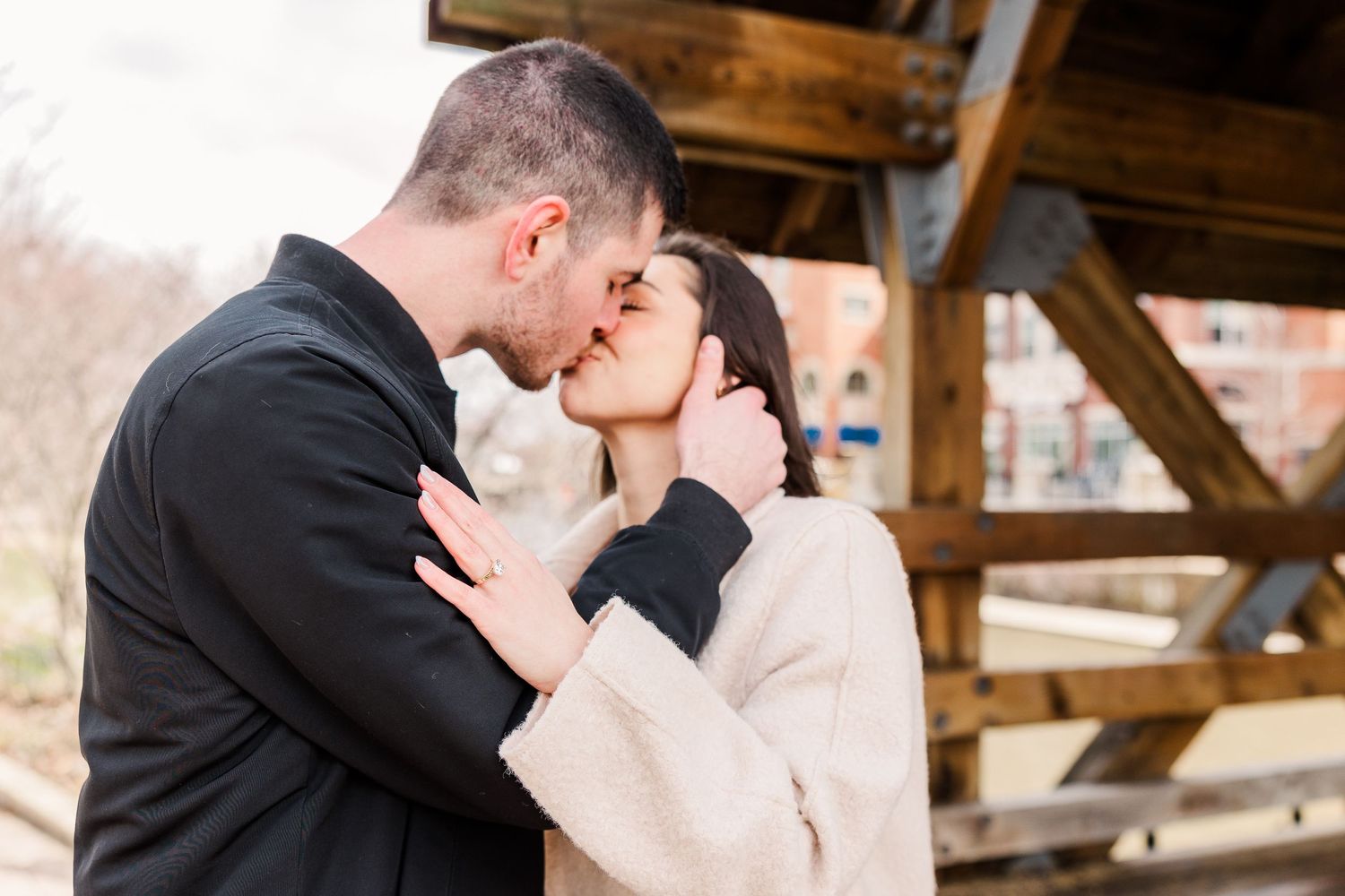 A couple shares a romantic kiss under a wooden structure outdoors.