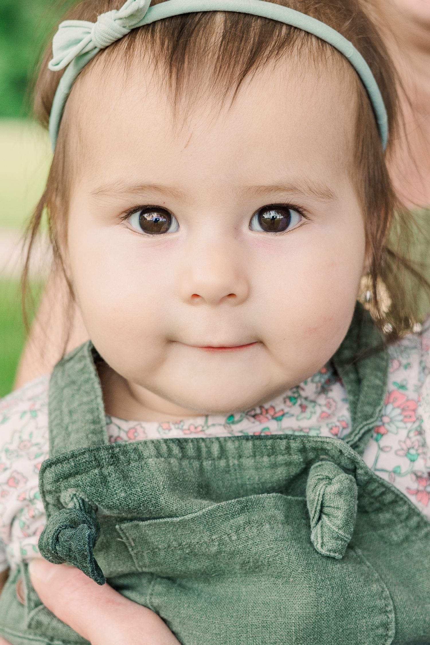 A sweet toddler wearing a mint green bow headband and overalls smiles at the camera.