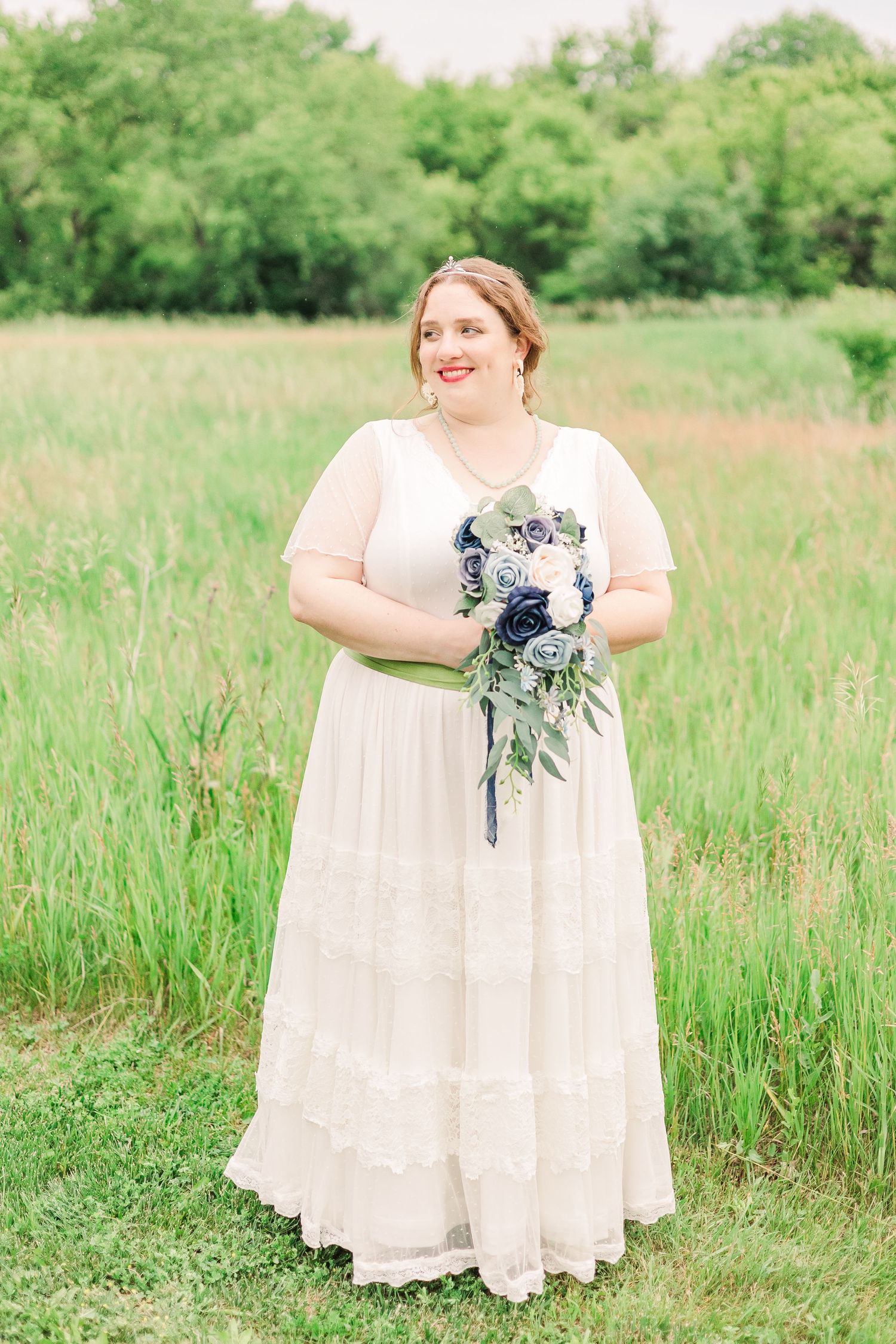 Bride in white dress with green sash holds blue and white bouquet while standing in meadow.