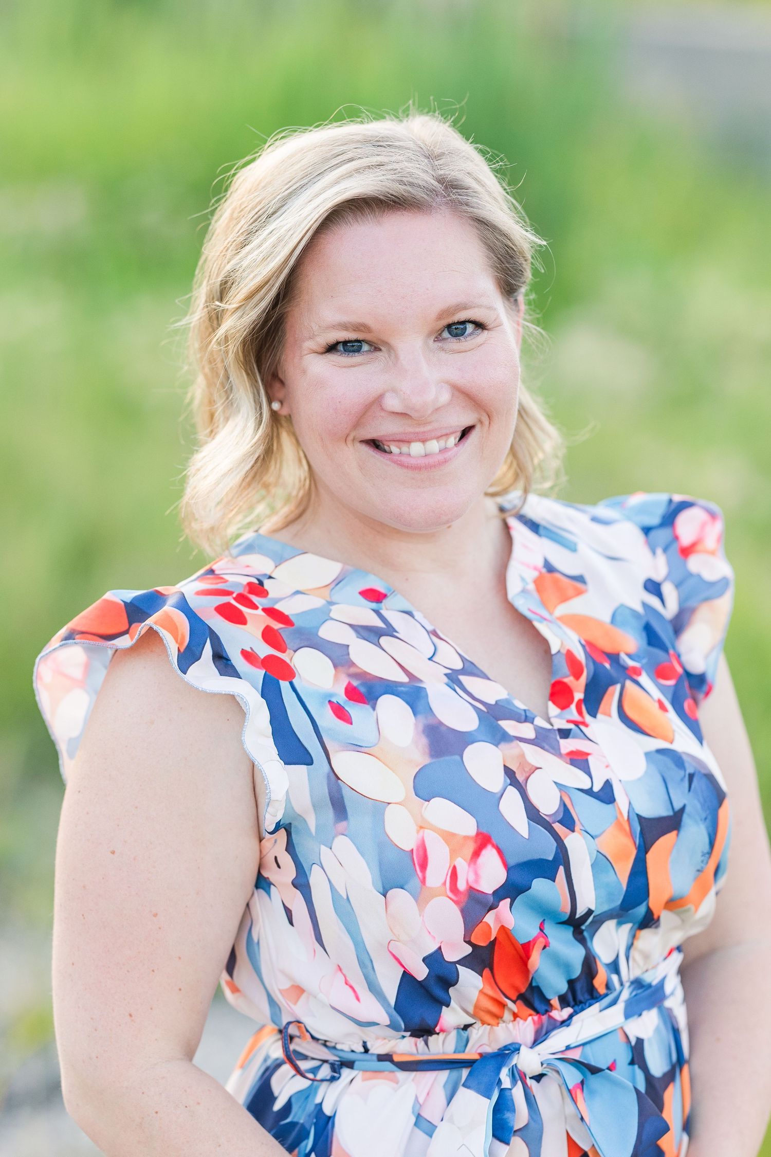 Woman in blue and coral floral print dress smiles outdoors in bright natural lighting.
