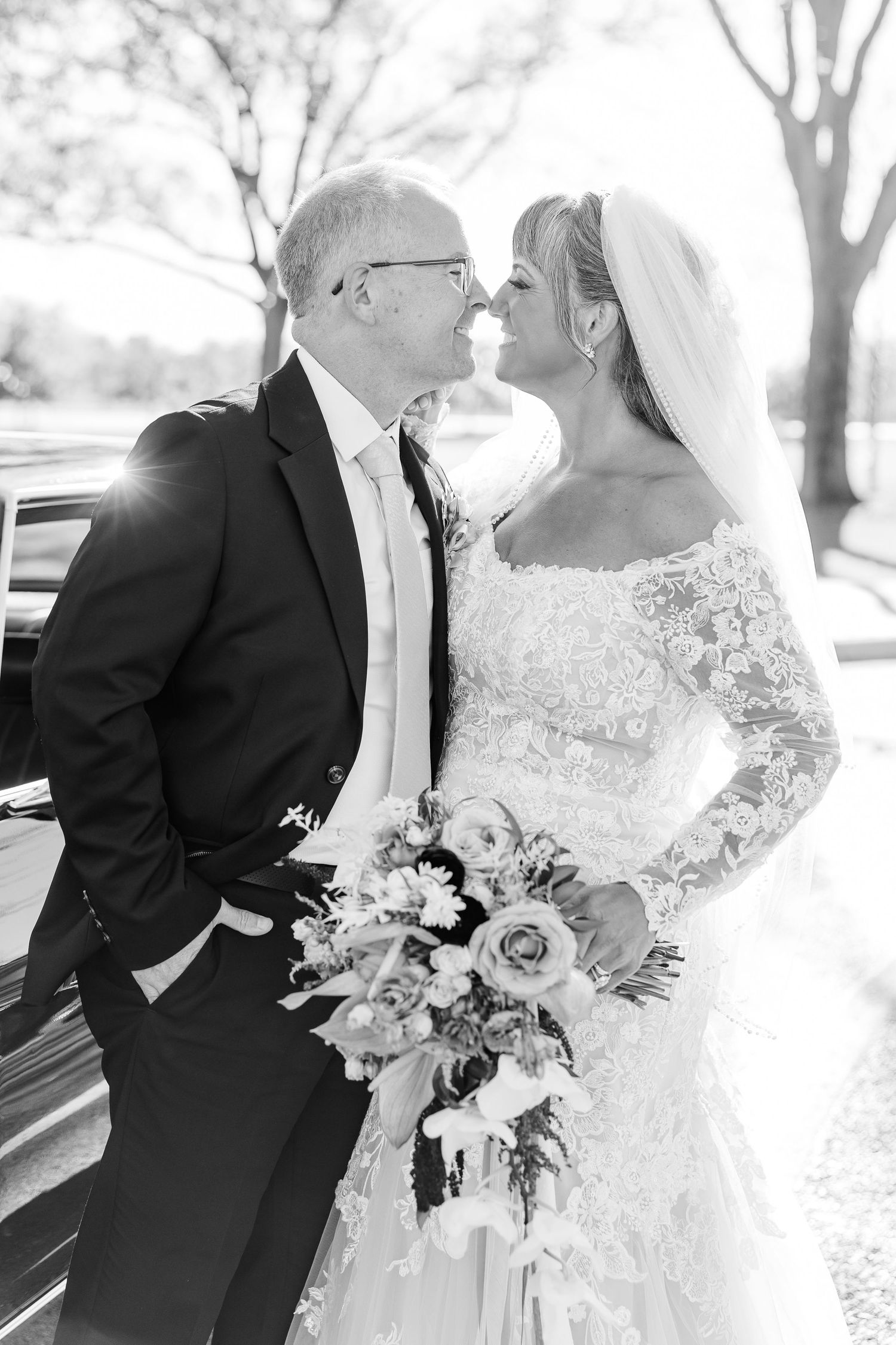 A black and white photo of a groom in a suit and a bride in a lace gown and veil nose to nose, holding a bouquet outdoors.