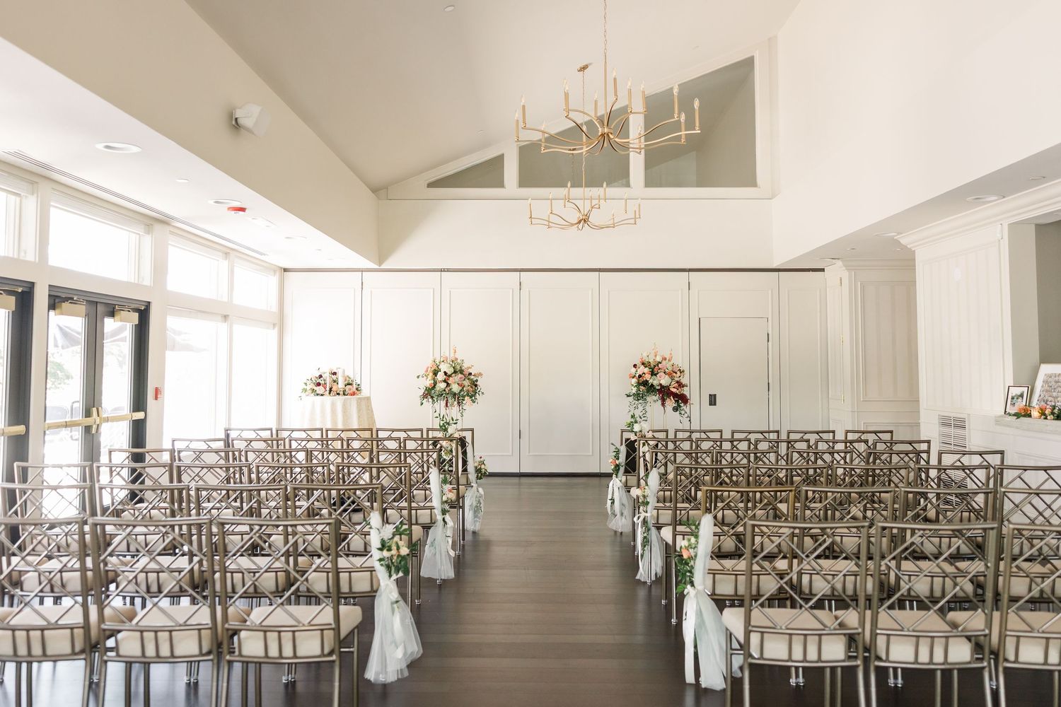 Bright indoor wedding ceremony venue with rows of gold chiavari chairs, white floral arrangements, a center aisle and a chandelier.
