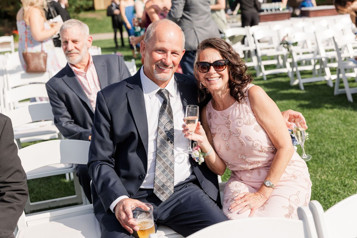 Smiling couple at outdoor wedding reception holding champagne and beer glasses.