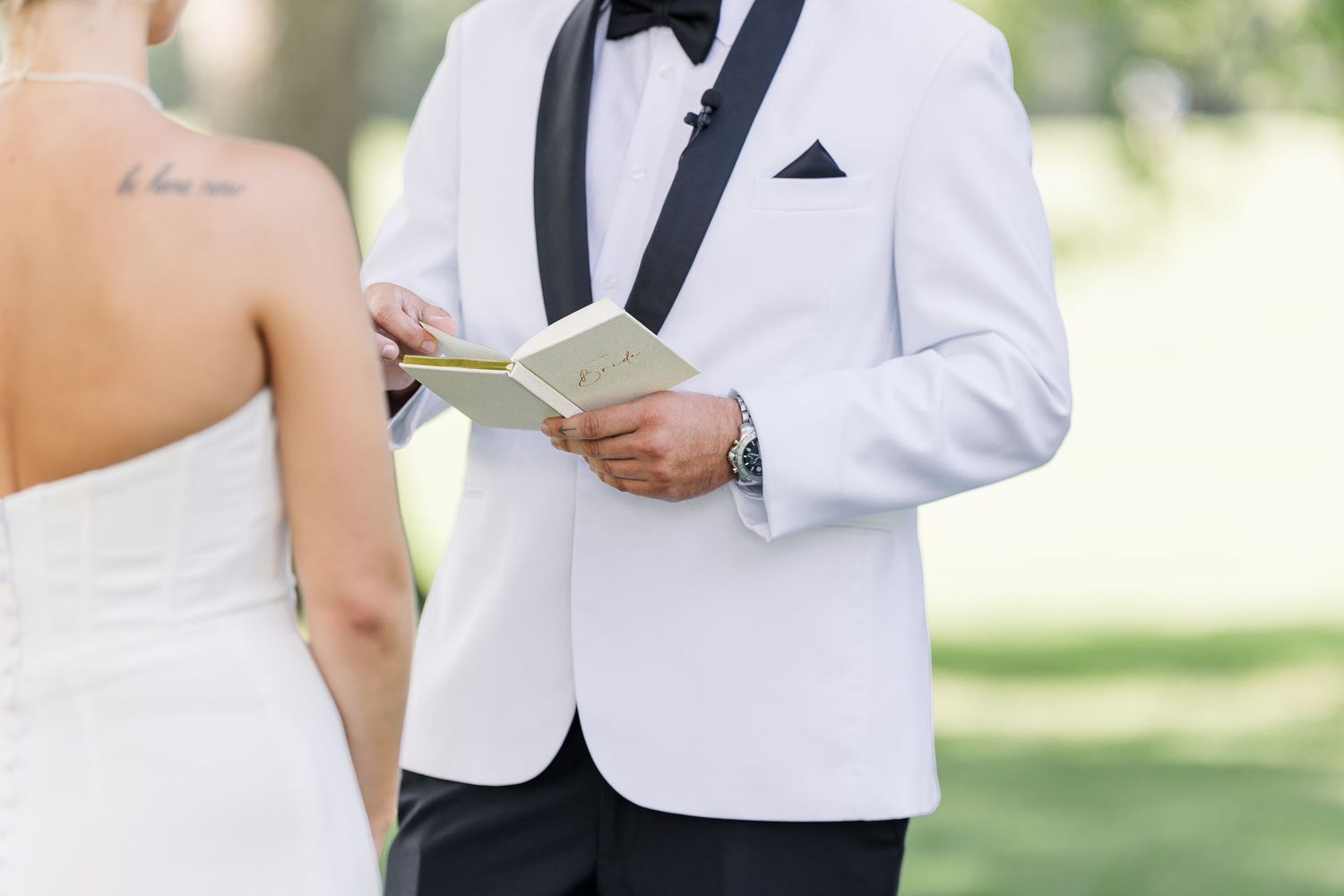 Groom in white tuxedo reads vows from booklet during outdoor wedding ceremony.