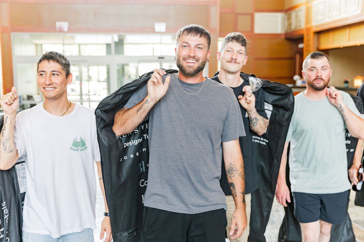 Group of five smiling men posing together holding black garment bags at an indoor venue.