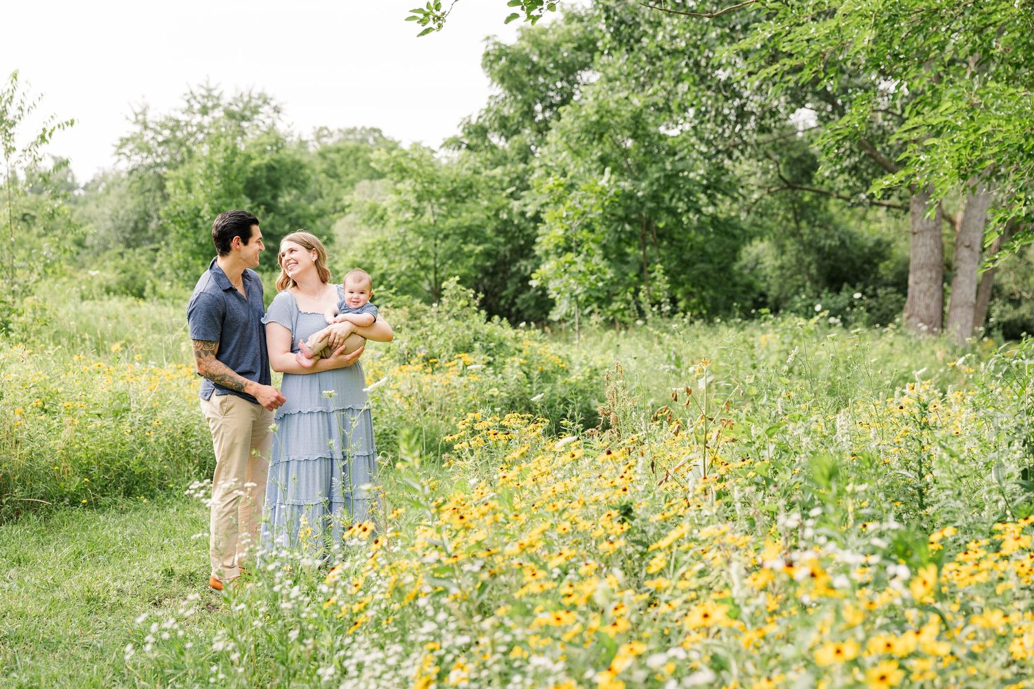 Family of three standing in a wildflower meadow with yellow blooms and lush green trees in summer.