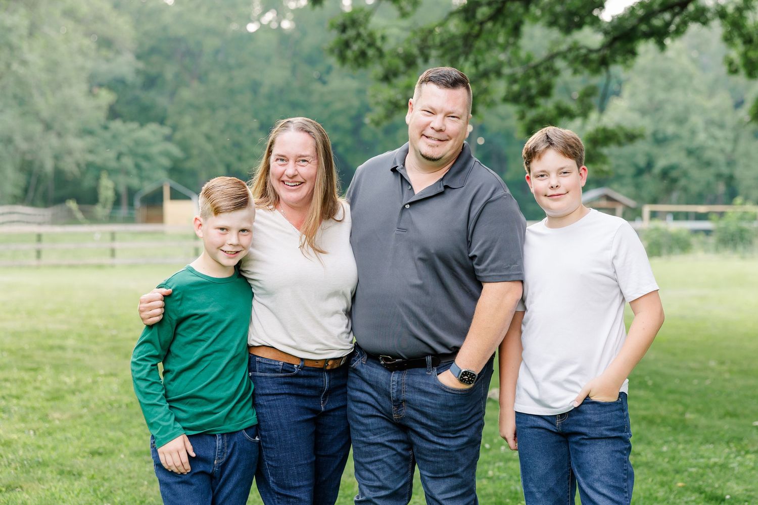 Family of four smiling outdoors on green grass with trees in background on a sunny day.