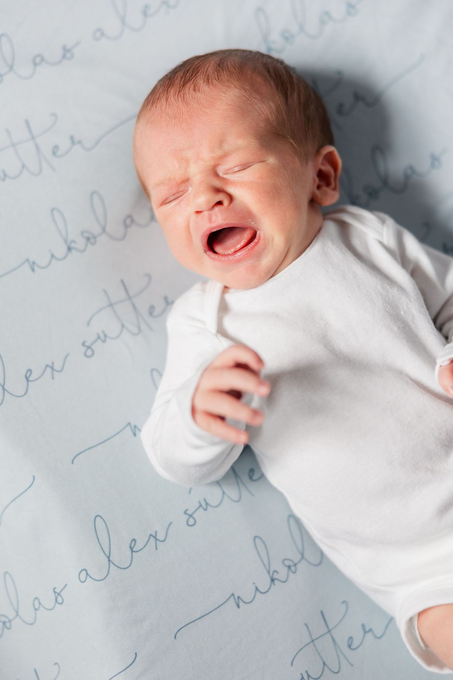 Crying newborn baby in white onesie lying on personalized blue blanket with cursive name print.