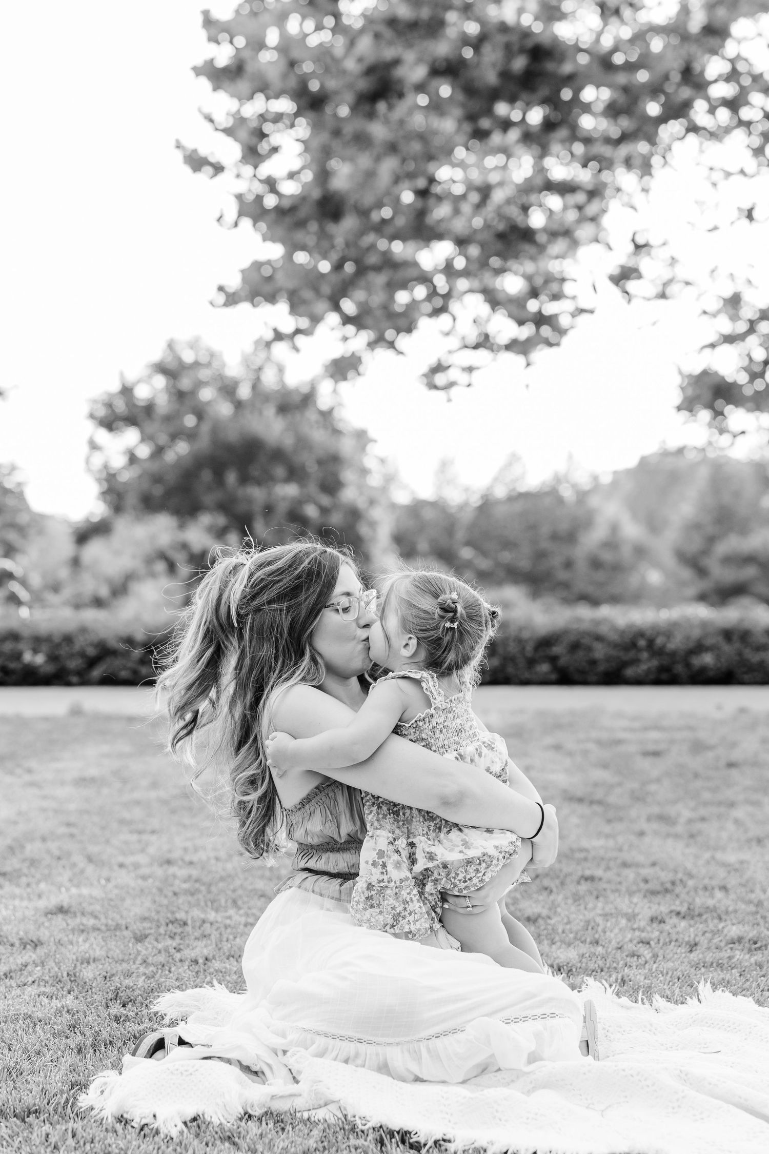 Black and white photo of mother sitting on grass kissing young daughter in tender outdoor moment.