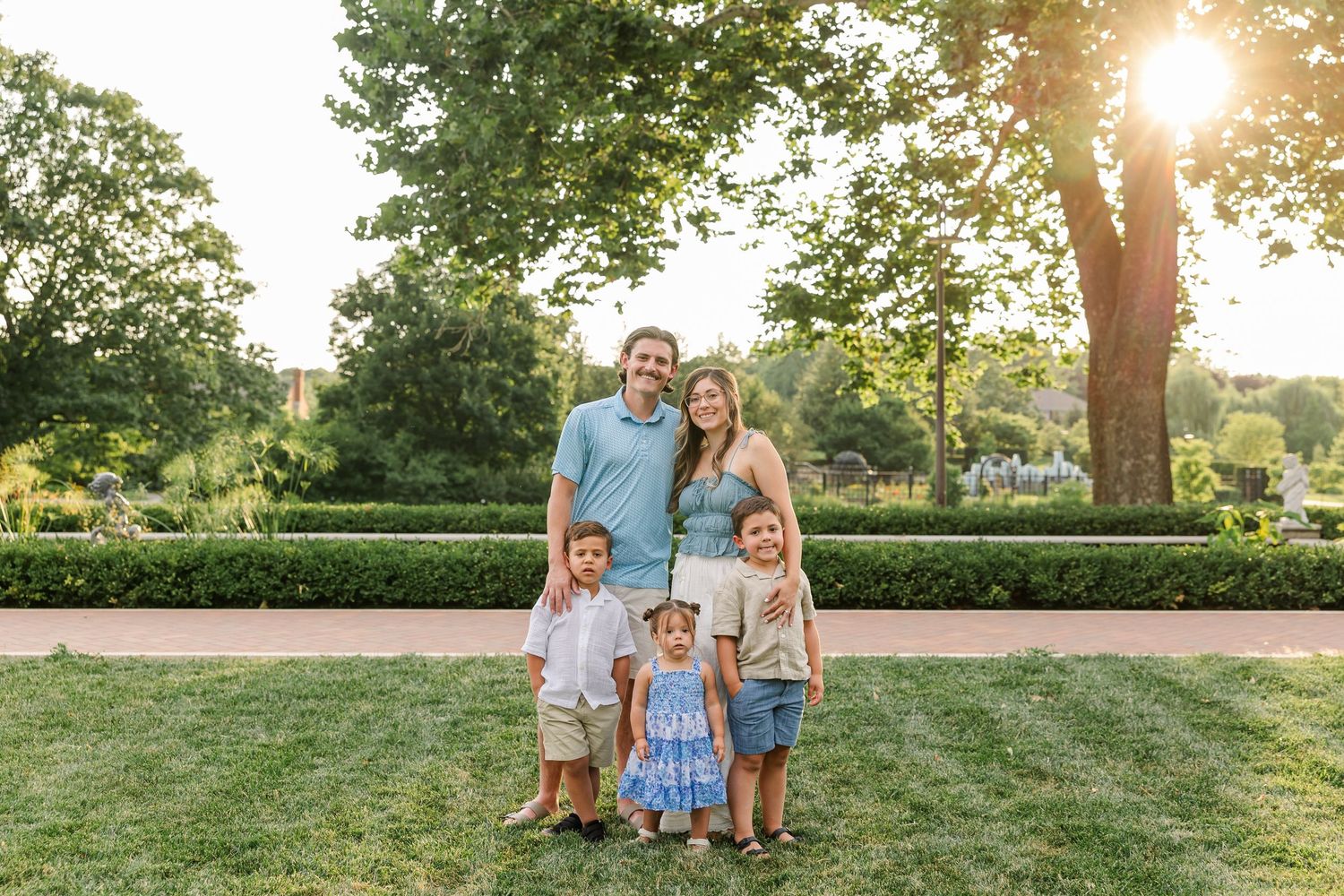Family of five posing outdoors on green lawn with lush trees and golden sunlight in background.