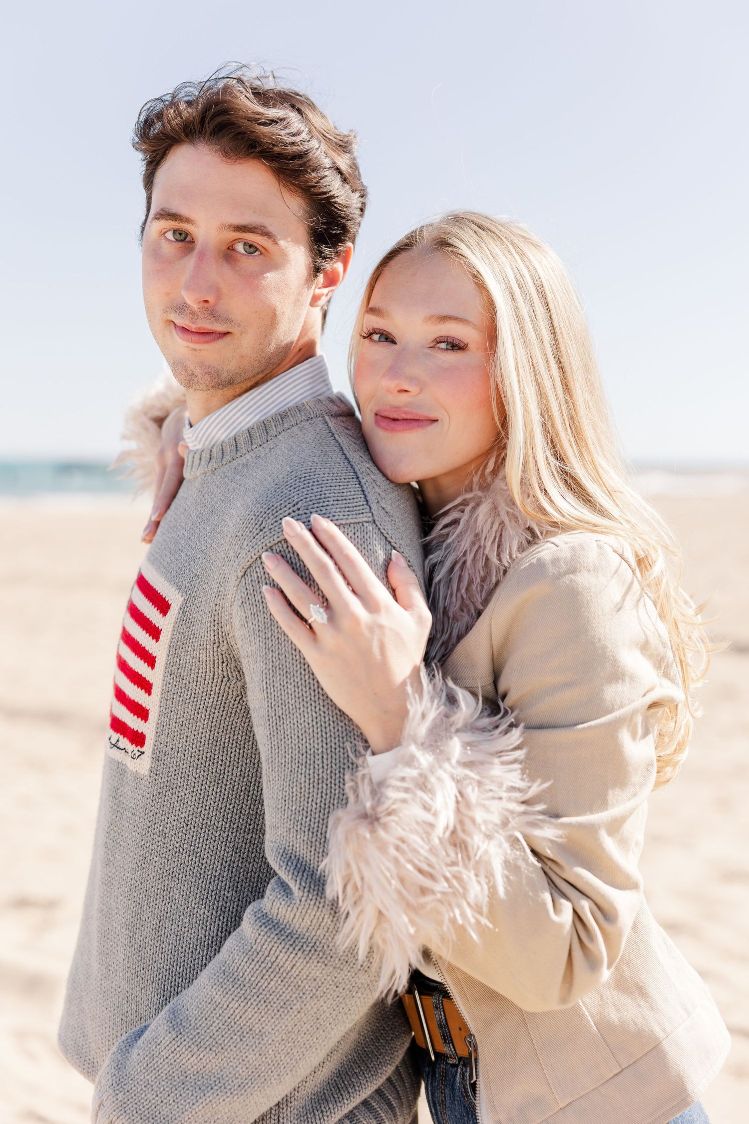 A couple embraces on a sunny beach, the man in a gray sweater and woman in a beige fur-trimmed jacket.