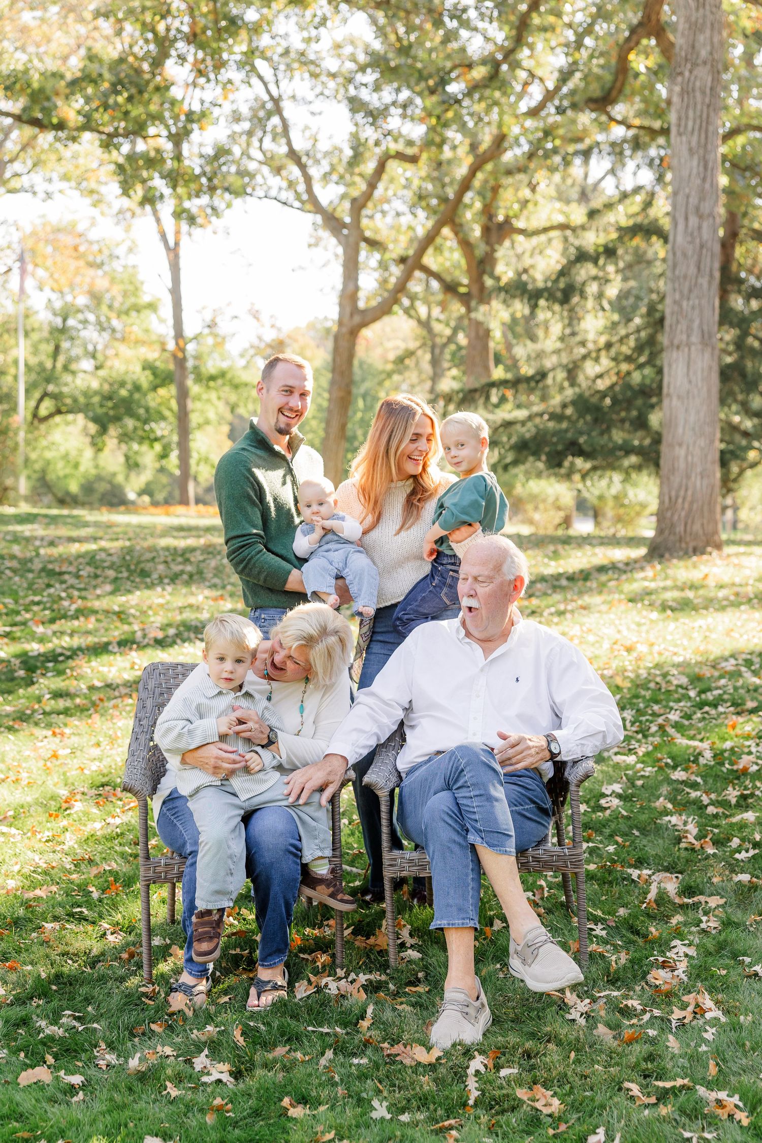 Multi-generational family portrait outdoors in a park, laughing together with two young children among autumn leaves.