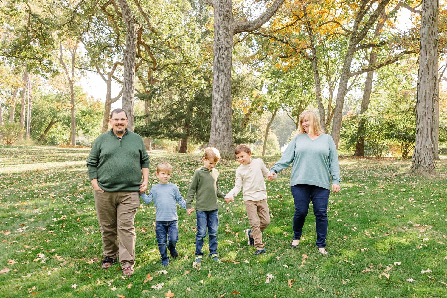 A happy family of five walks hand-in-hand through a tree-lined park during autumn foliage season.
