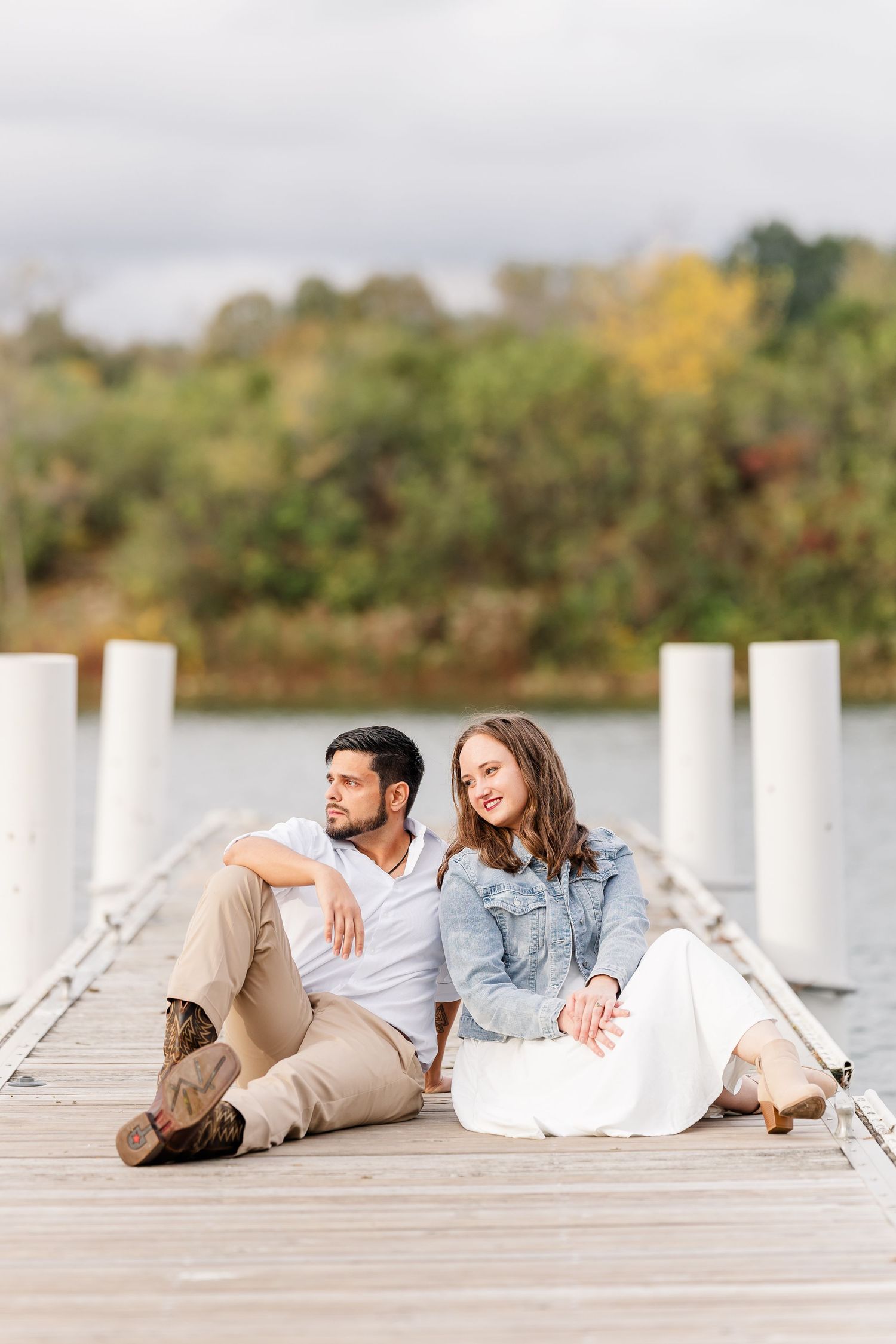 Couple sitting close together on a wooden lakeside dock with white posts, autumn trees and a cloudy sky behind them.