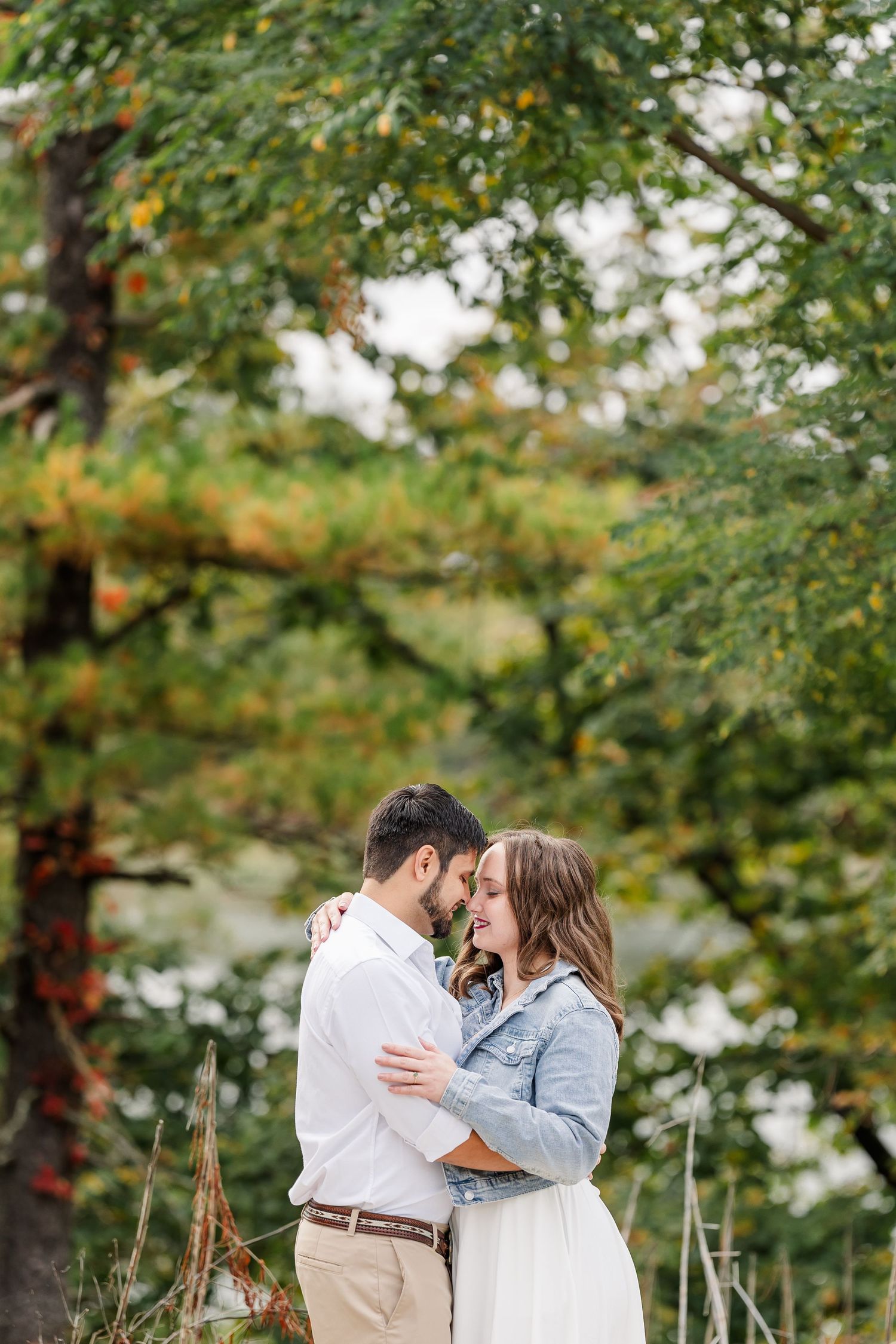 Couple embracing and smiling in a park surrounded by colorful autumn trees during an outdoor engagement photo session.