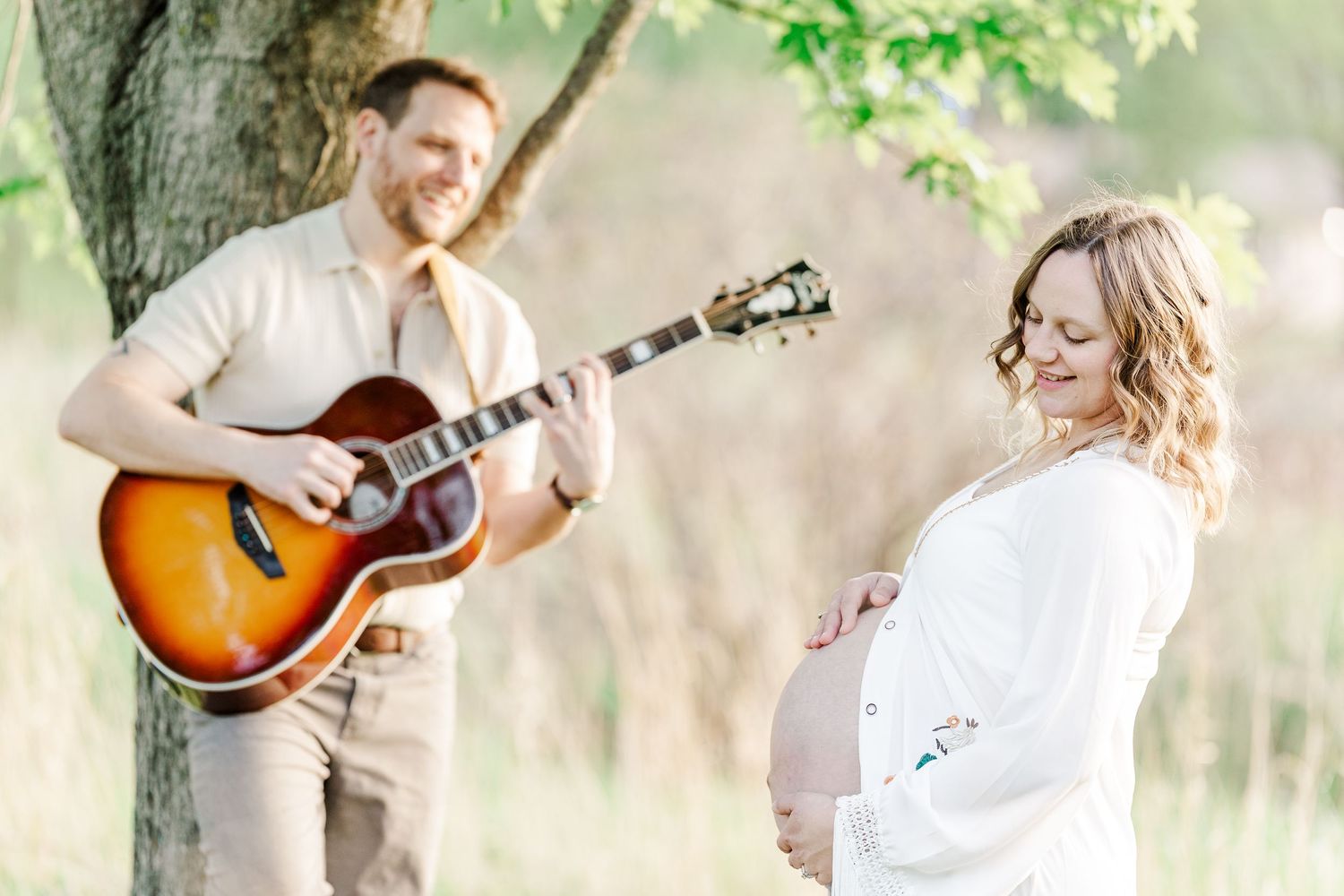 Pregnant woman in white dress stands outdoors while man plays acoustic guitar beneath a tree during maternity photo session.