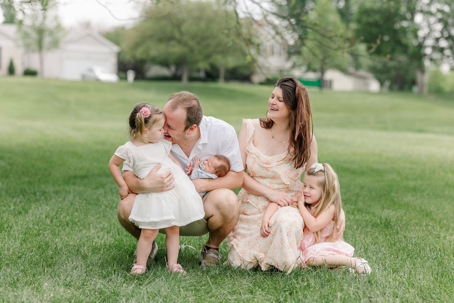 Happy family of five with young children and newborn baby laughing together on lush green lawn.