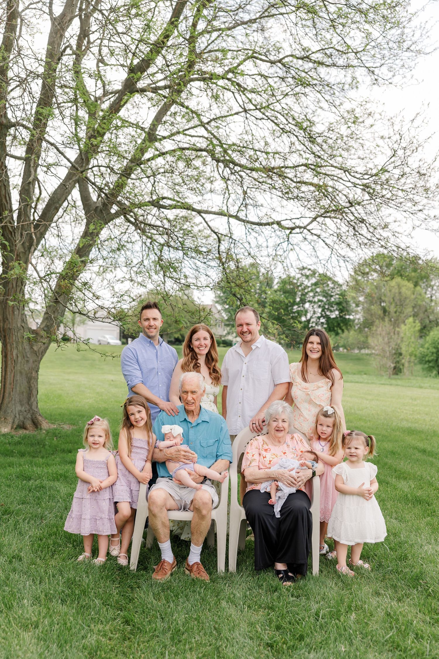 Multi-generational family portrait with grandparents seated and adult children and grandkids posing outdoors under a tree.