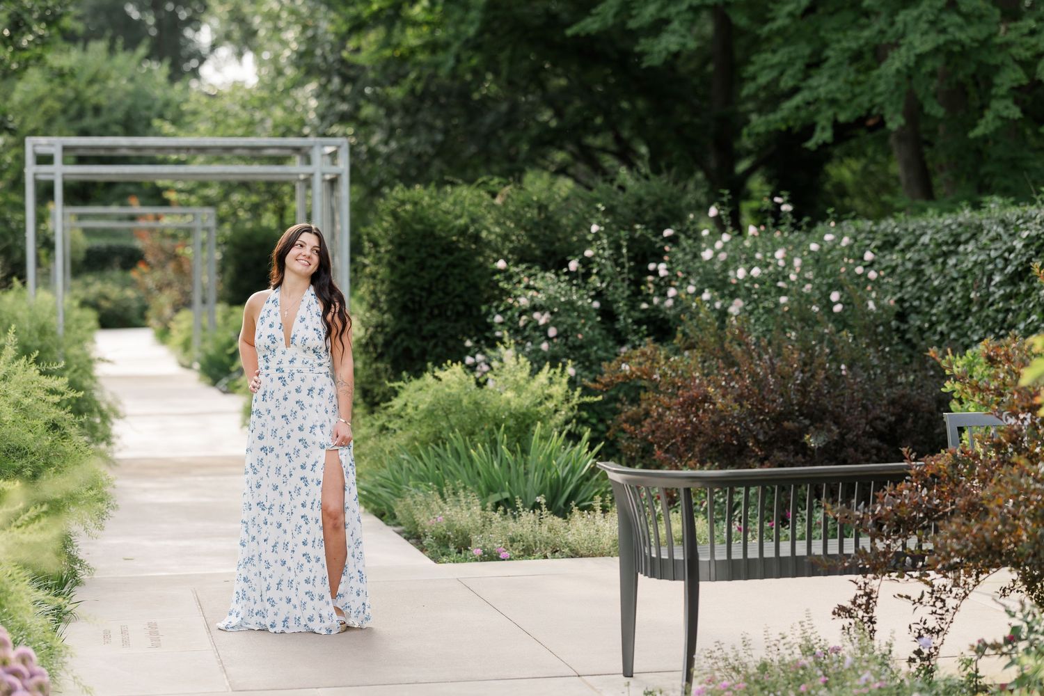 Woman in floral maxi dress posing in a lush garden pathway with greenery and white flowers.