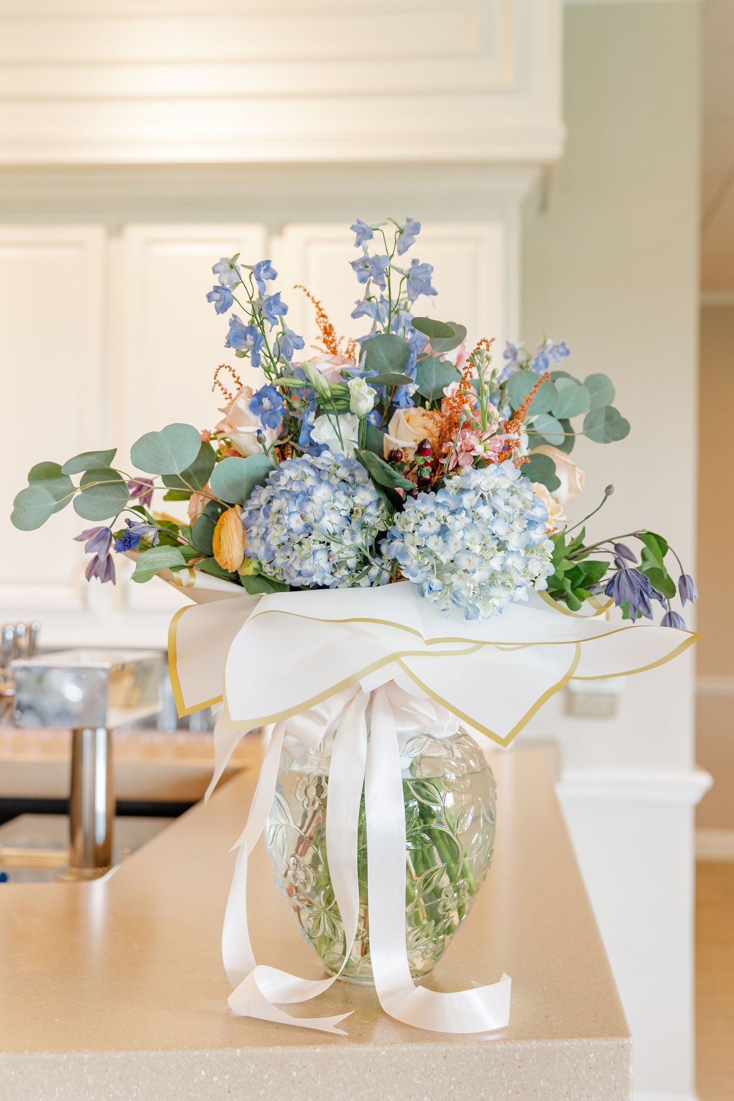 Elegant floral arrangement with blue hydrangeas and mixed blooms in a glass vase with white ribbon bow on kitchen counter.