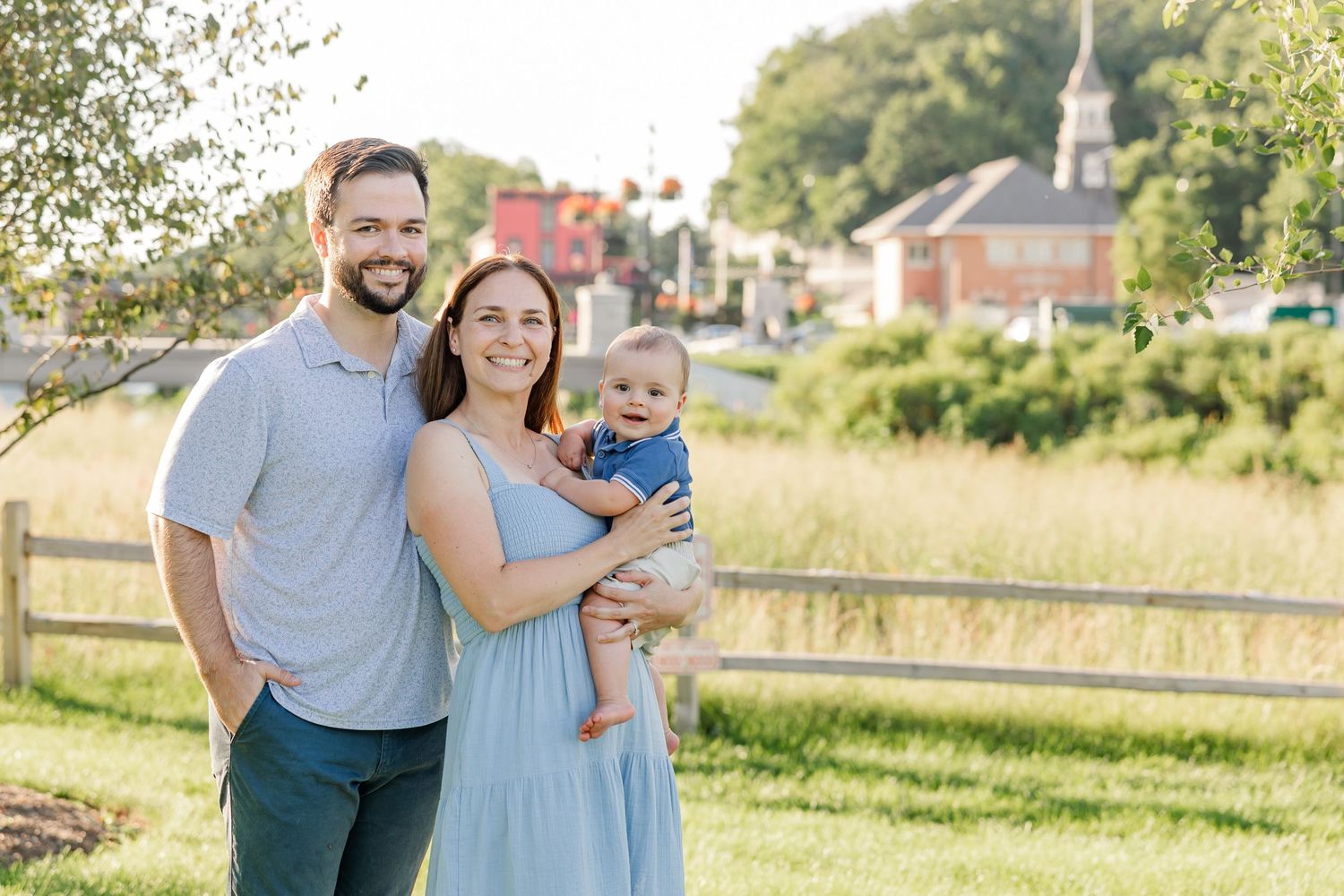 A smiling couple stands outdoors holding their baby boy, with a scenic New England town background.