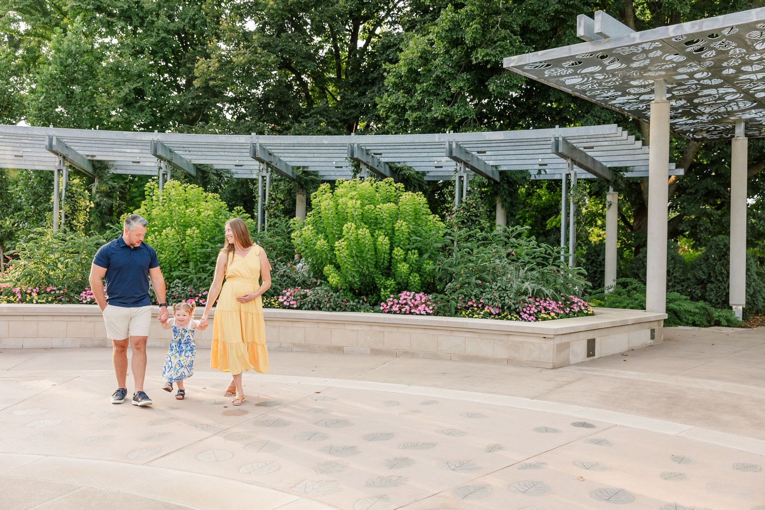 Family of three holding hands walking past colorful garden beds near modern pergola structure.