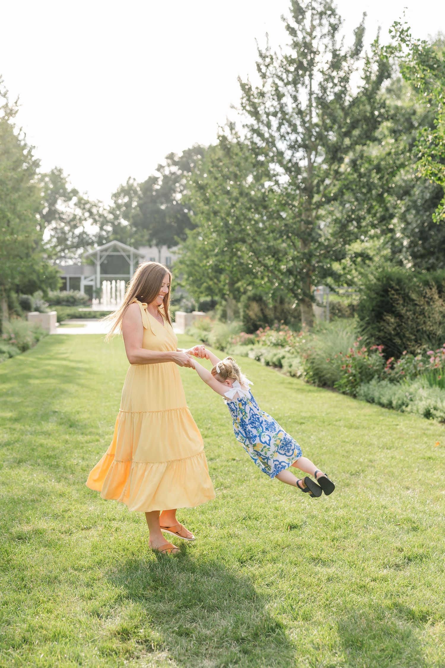 Mother in yellow dress spinning laughing toddler girl on green lawn.