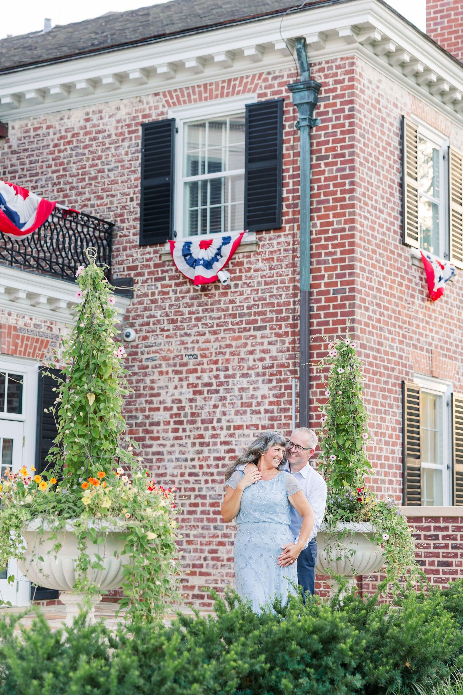 Romantic couple embracing outdoors by historic brick building adorned with patriotic bunting and lush garden planters.