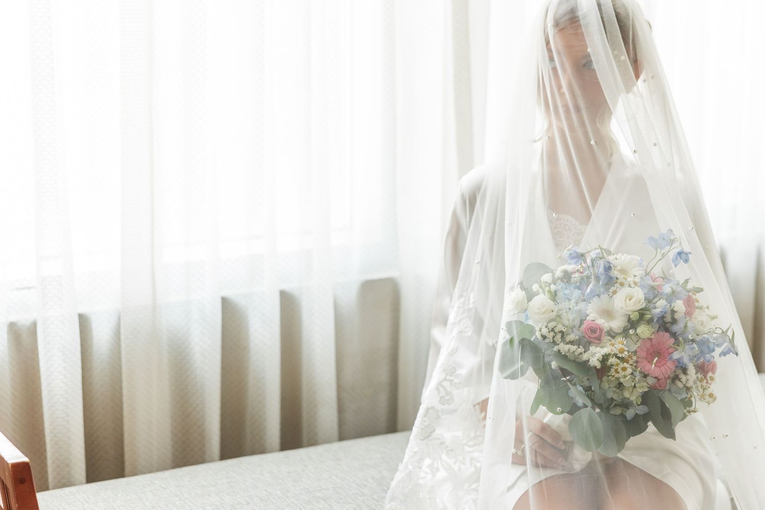 Bride in white lace veil holding a pastel bouquet of pink, blue, and white flowers, seated by a bright window.