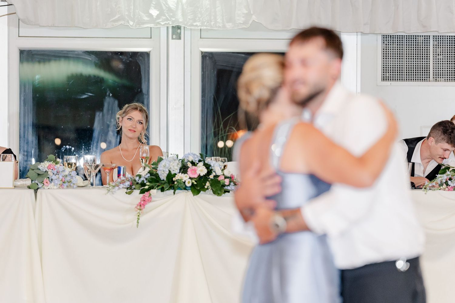 Bride watches emotionally as groom shares a dance with a guest during wedding reception at elegantly decorated venue.