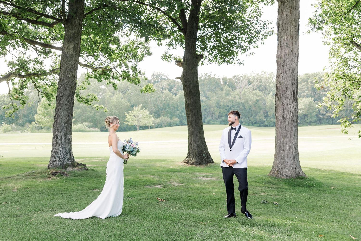Bride and groom share emotional first look moment outdoors on lush green lawn beneath tall trees.