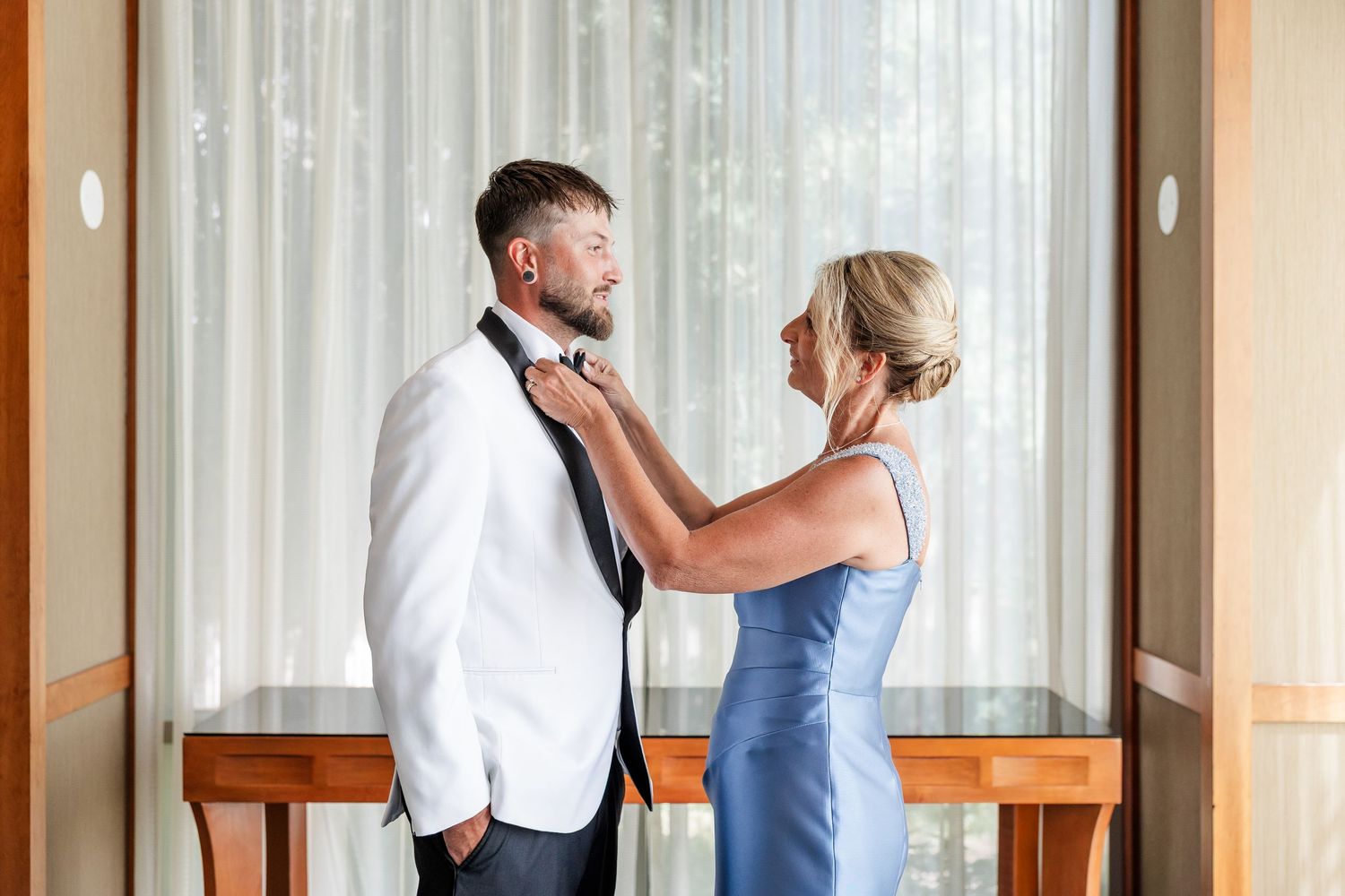 A woman in a blue dress adjusts the groom's black tie as he stands in a white tuxedo jacket by a window.