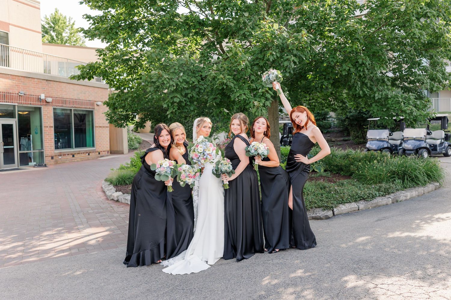 Bride and five bridesmaids in black gowns laugh and pose together outdoors on summer wedding day.