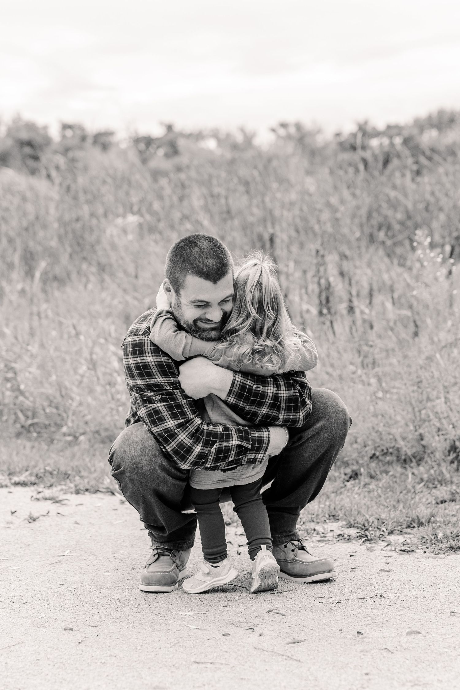 Black and white photo of a bearded dad crouching to embrace his young daughter on an outdoor path surrounded by tall grass.